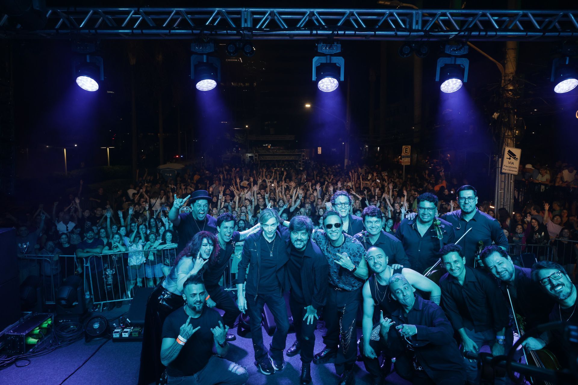 A banda posa para fotos com o público em um show ao ar livre; os músicos vestem roupas escuras e fazem gestos com as mãos.