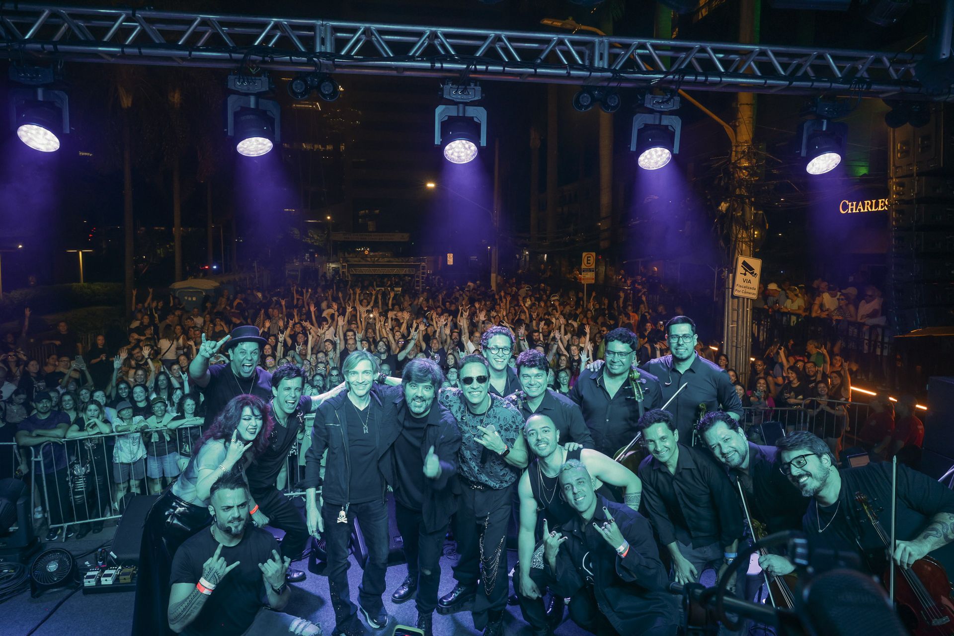 A banda posa para fotos com o público em um show ao ar livre; os músicos vestem roupas escuras e fazem gestos com as mãos.