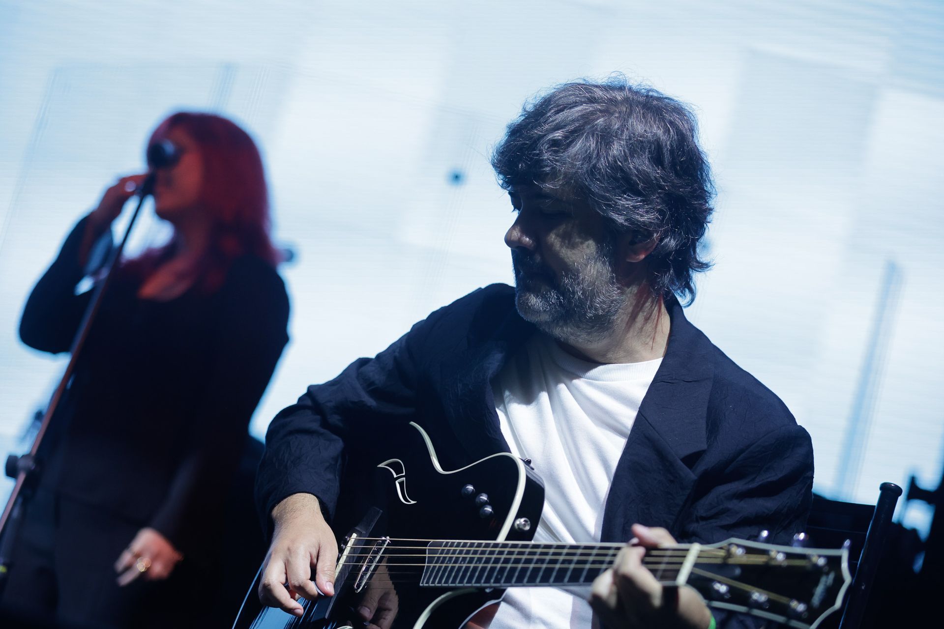 Um artista tocando violão e cantando, enquanto mulher canta ao microfone no palco.