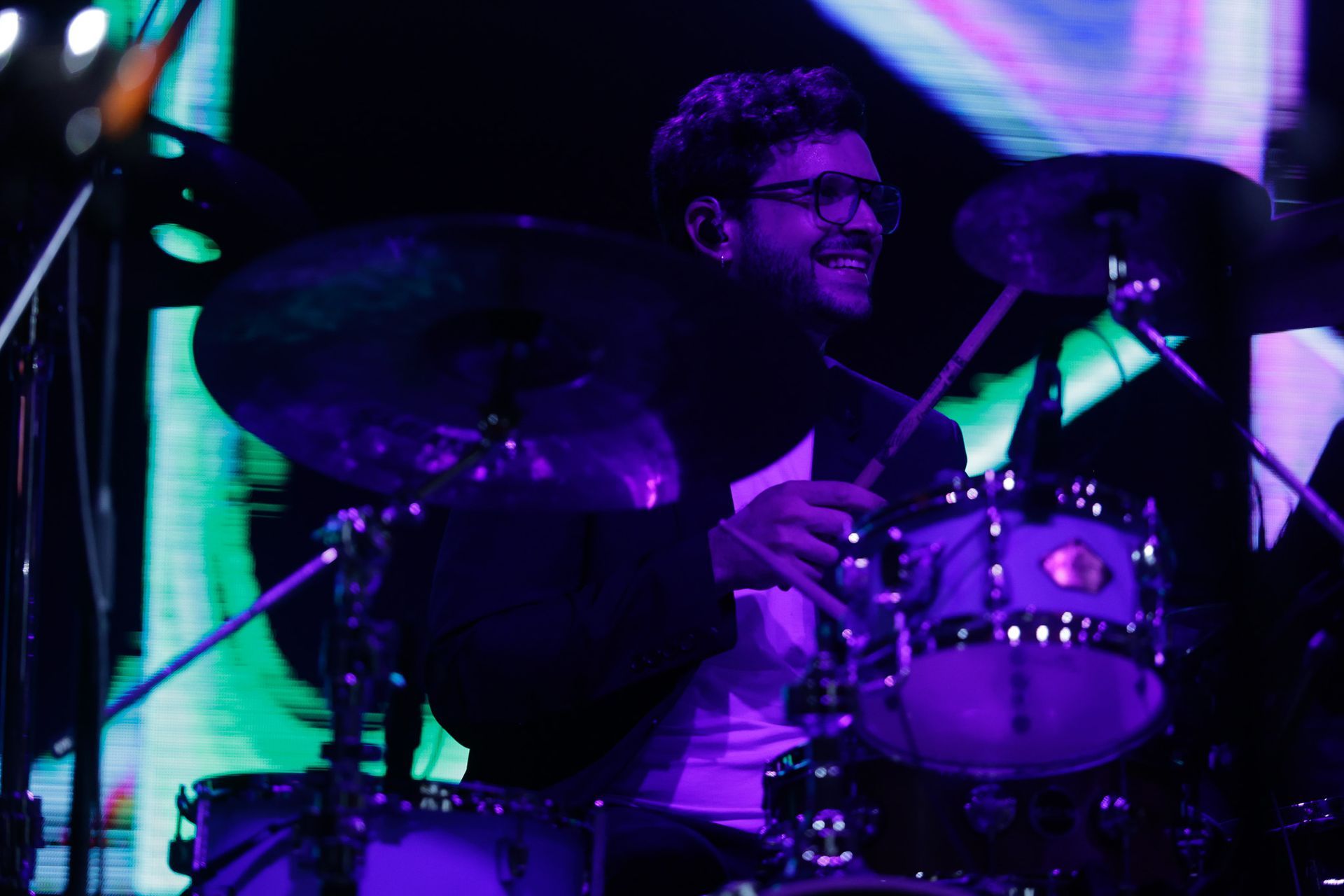 Baterista de óculos sorrindo, tocando bateria sob iluminação roxa no palco.