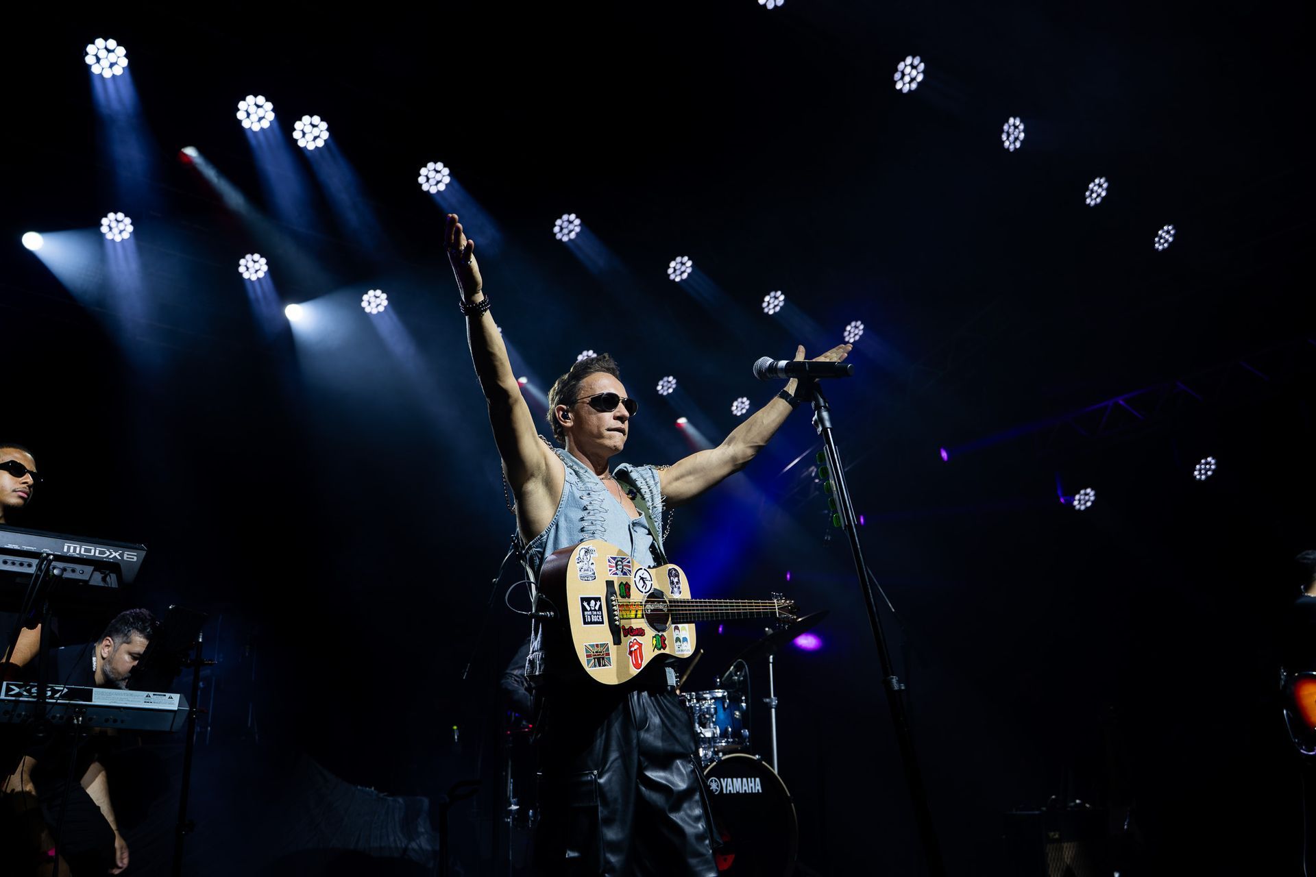 Um artista toca um violão acústico coberto de adesivos com os braços erguidos sob luzes de palco brilhantes.