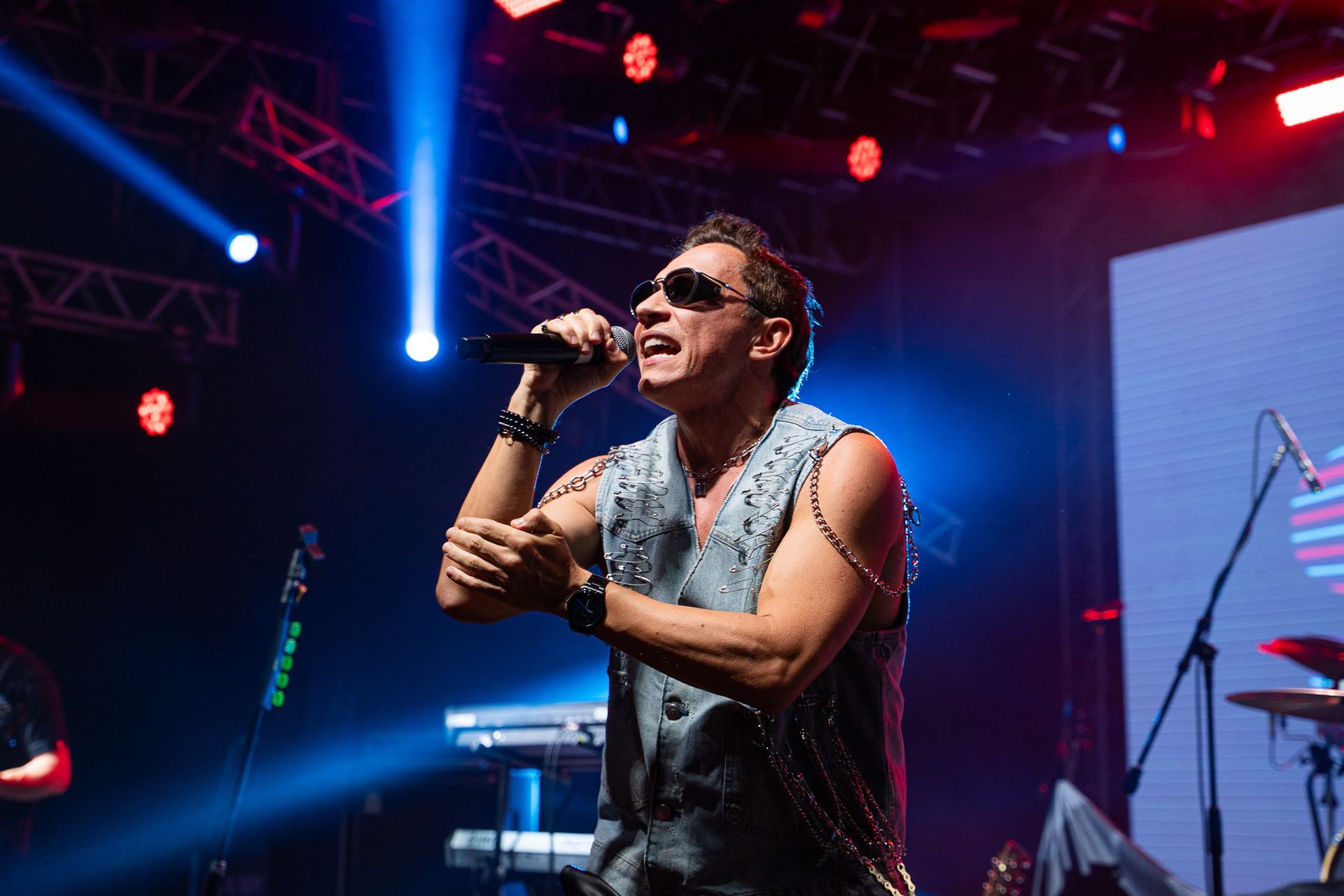 Vocalista de colete jeans e óculos escuros se apresentando no palco sob luzes azuis brilhantes.