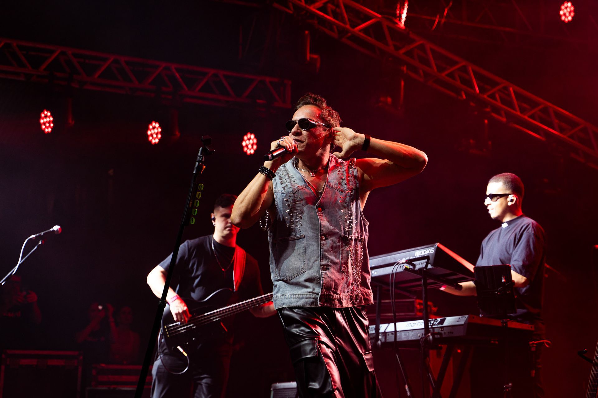 O vocalista principal se apresenta no palco com um guitarrista e um tecladista sob holofotes vermelhos.