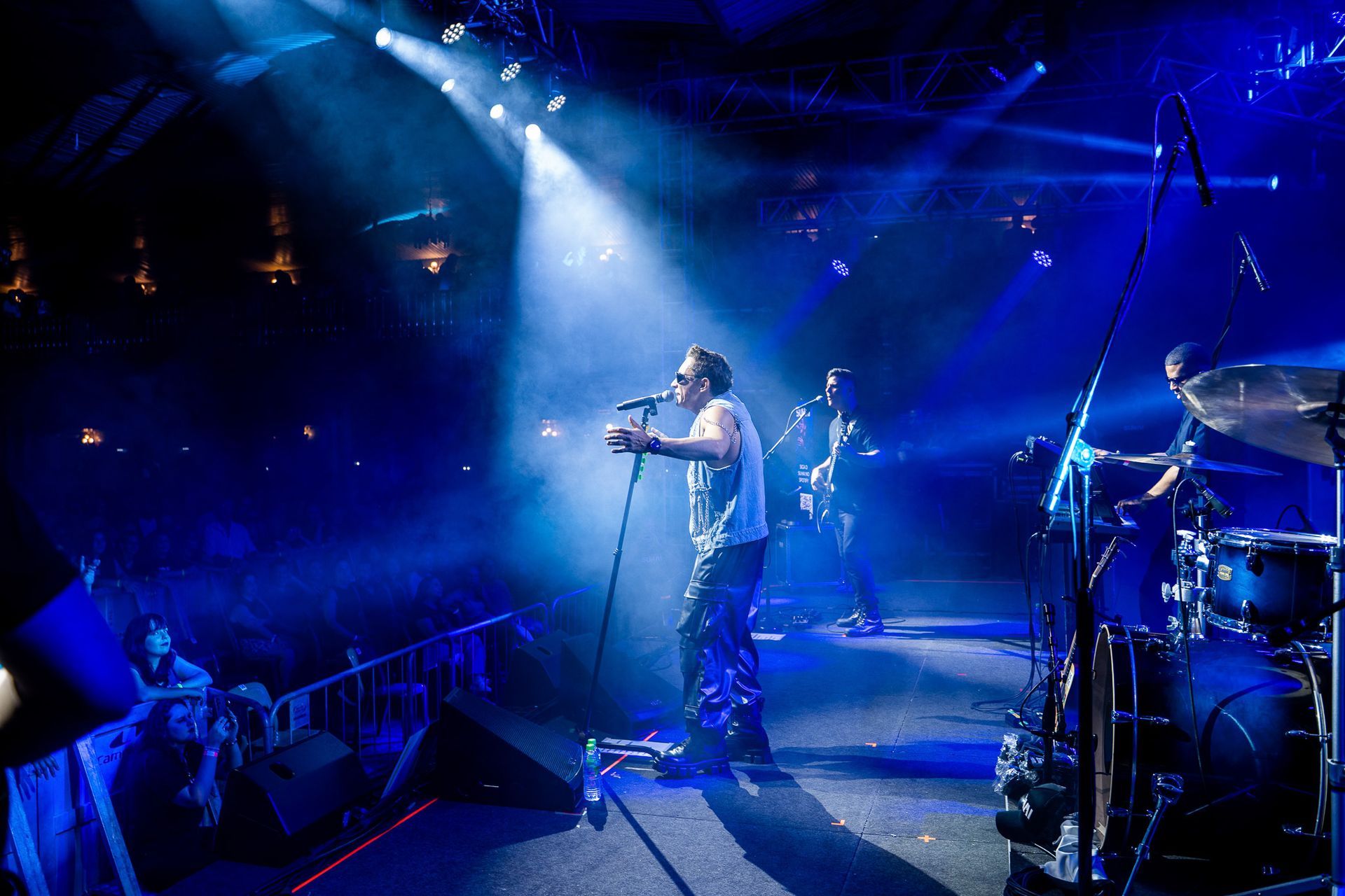 Um cantor se apresenta em um palco acompanhada por um baterista e um baixista em uma sala de concertos com pouca luz.