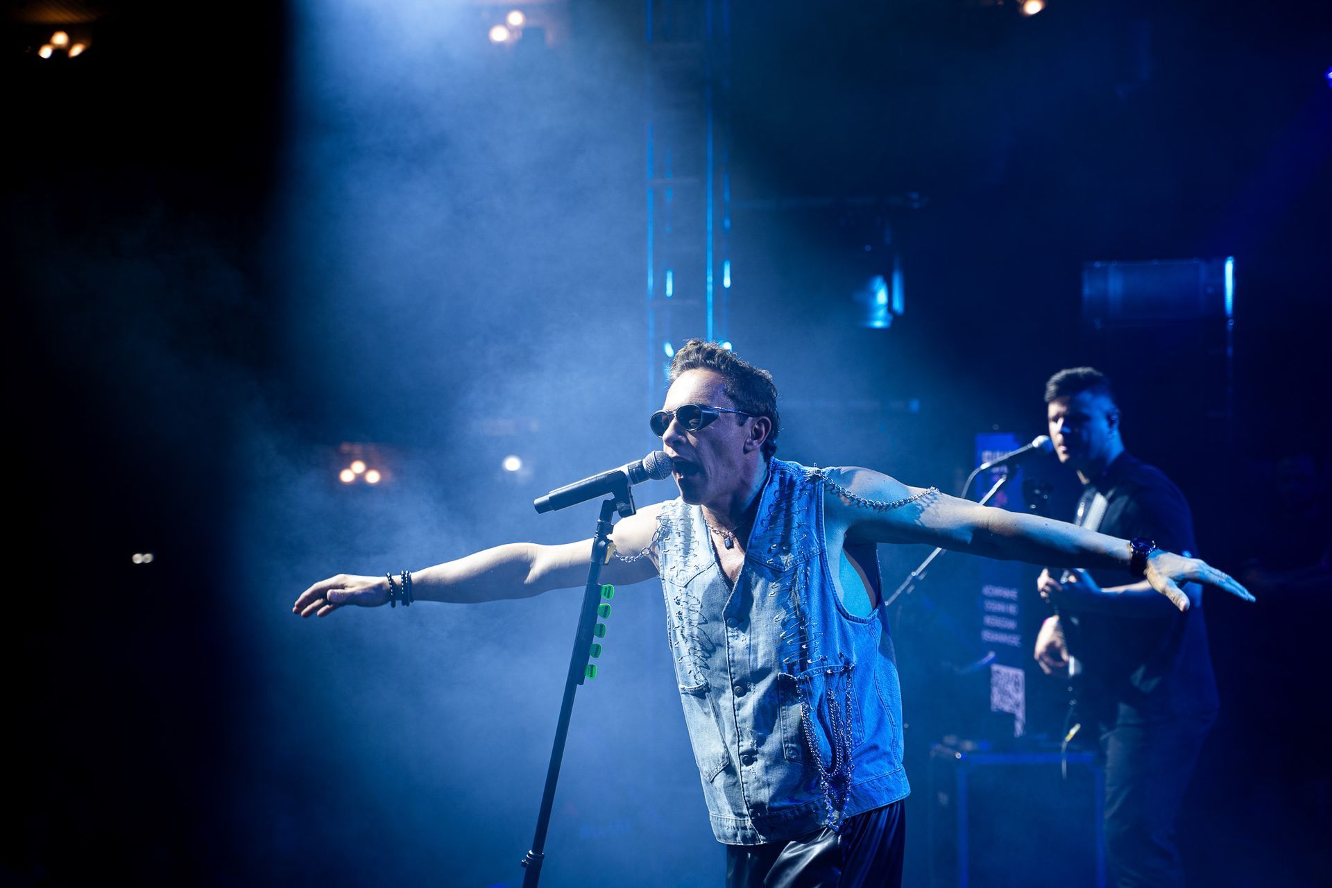 Um músico com um colete cravejado de tachas canta ao microfone no palco