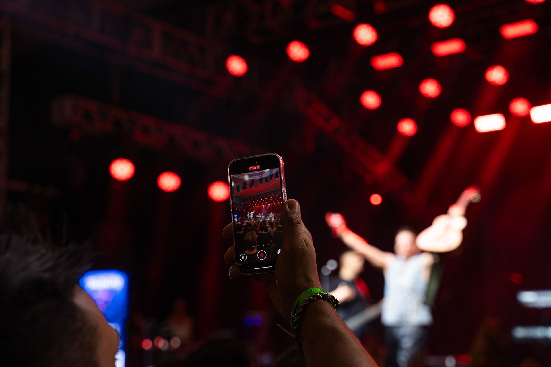 Uma mão segura um smartphone para gravar um músico em um palco iluminado por luzes vermelhas de concerto.