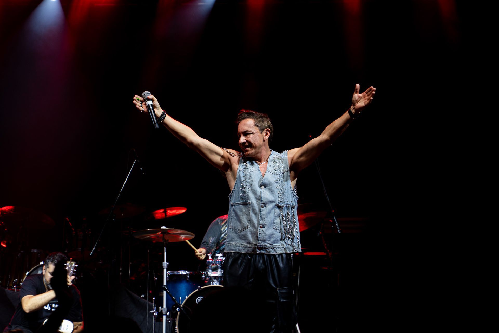 Um artista em um palco escuro de concerto, com os braços erguidos, vestindo um colete jeans sem mangas