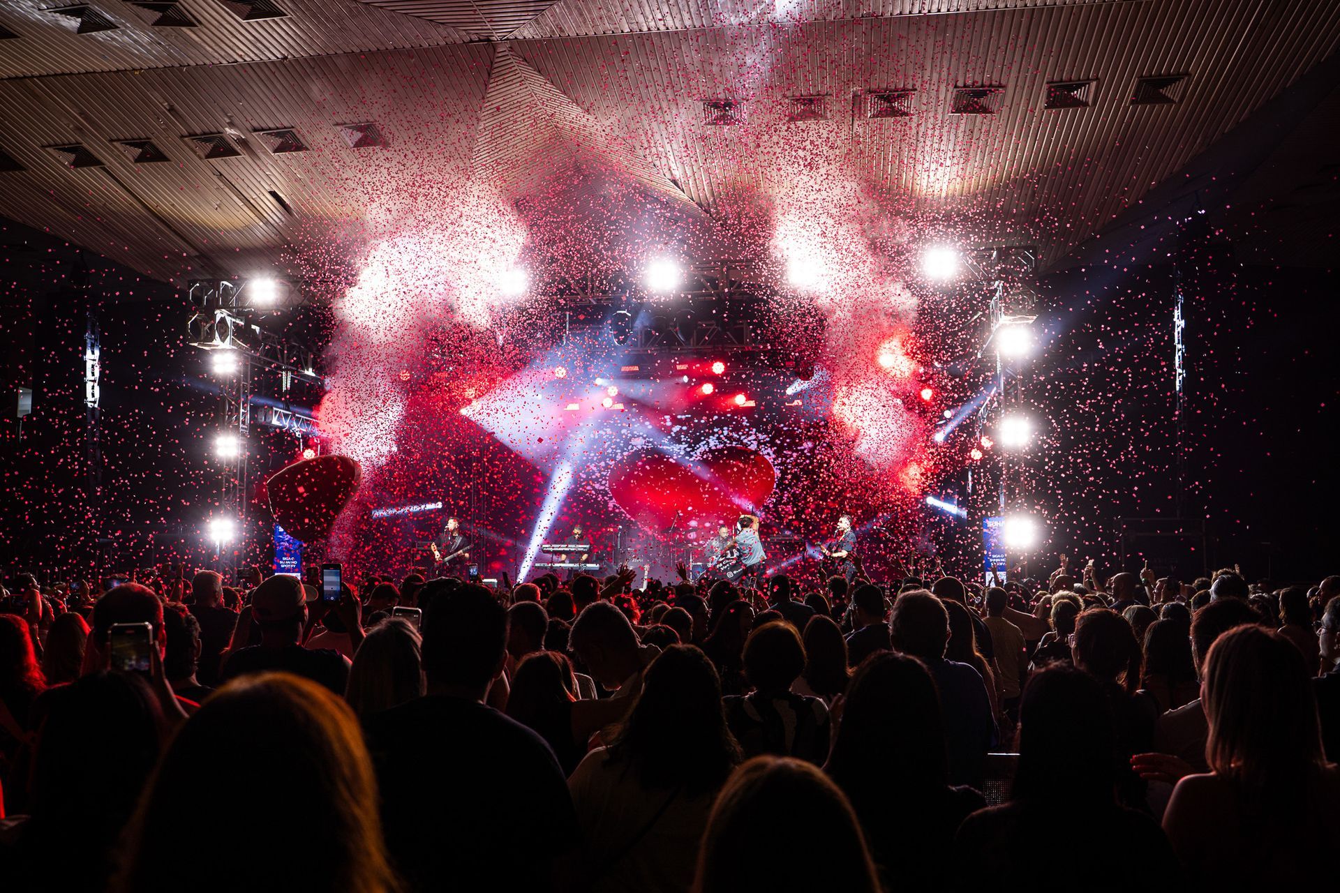 Uma multidão assiste a uma apresentação no palco, com decorações vermelhas em forma de coração e confetes 