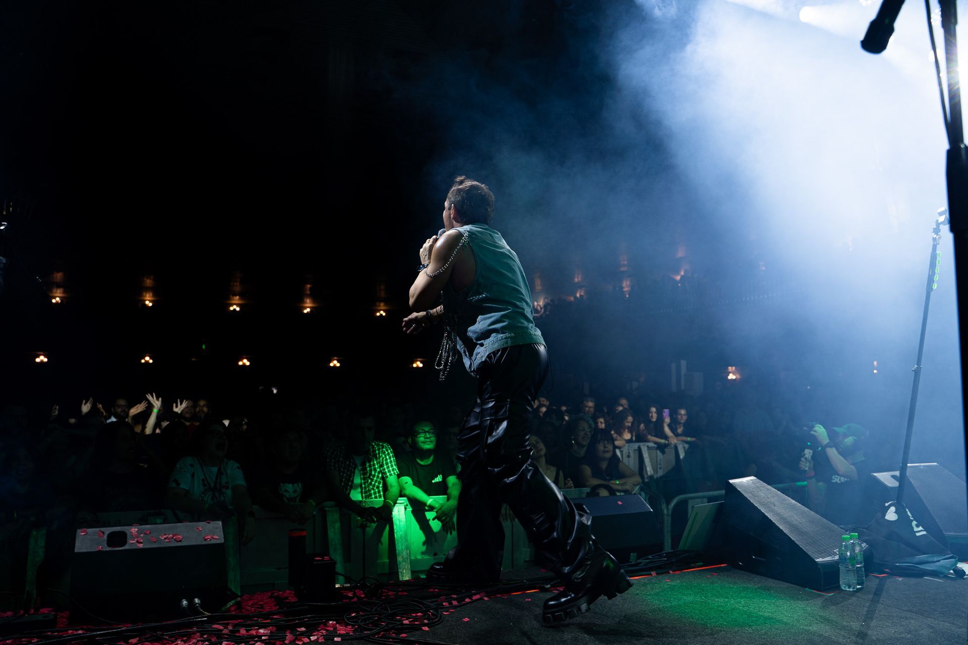 Um artista no palco cantando ao microfone sob luzes azuis, de frente para uma plateia em silhueta à noite.
