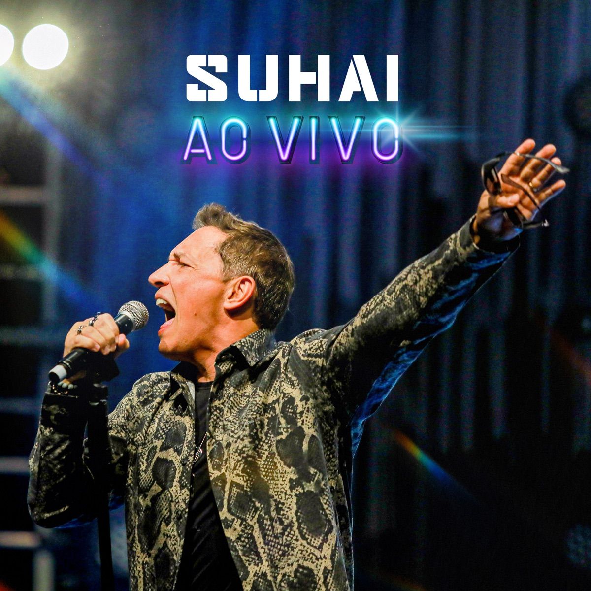 Cantor segurando um microfone no palco com a inscrição “Suhai Ao Vivo” e luzes azul-púrpura ao fundo.