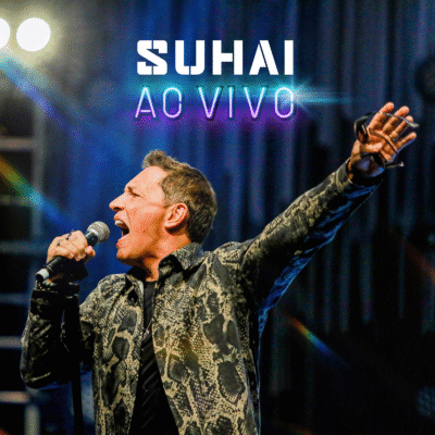 O cantor Suhai se apresenta ao vivo, segurando o microfone, com o braço levantado. Luzes de palco, texto 