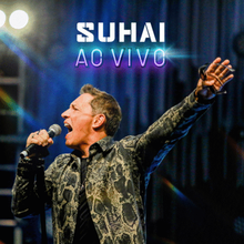 O cantor Suhai se apresenta ao vivo, segurando o microfone, com o braço levantado. Luzes de palco, texto 