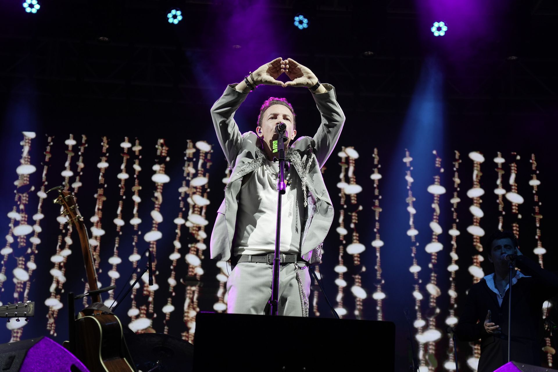 Cantor no palco com os braços erguidos, os dedos formando um coração. Luzes de palco roxas e brancas.