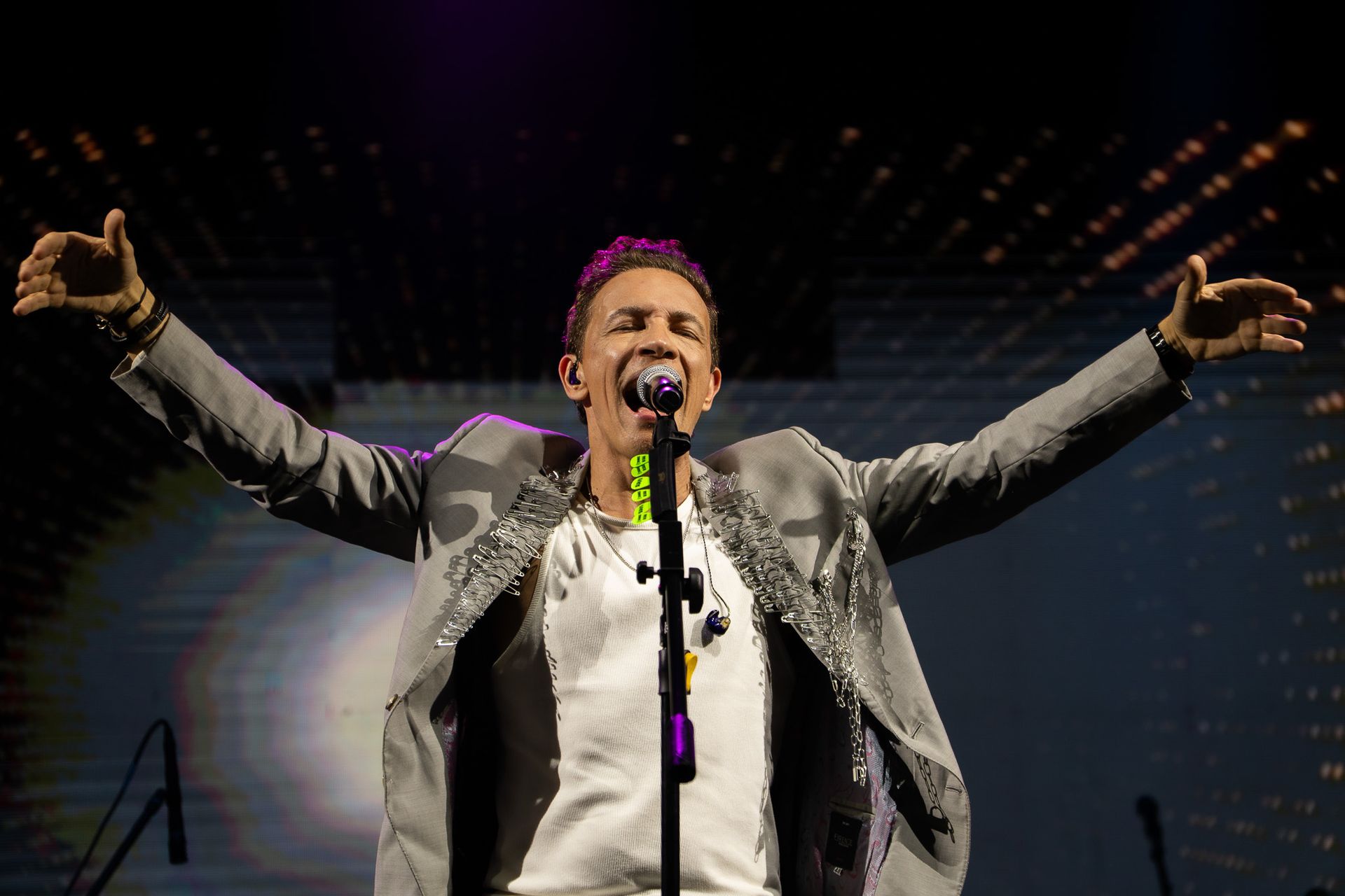 Um artista cantando em um microfone no palco, com os braços estendidos, vestindo uma jaqueta cinza com detalhes prateados.
