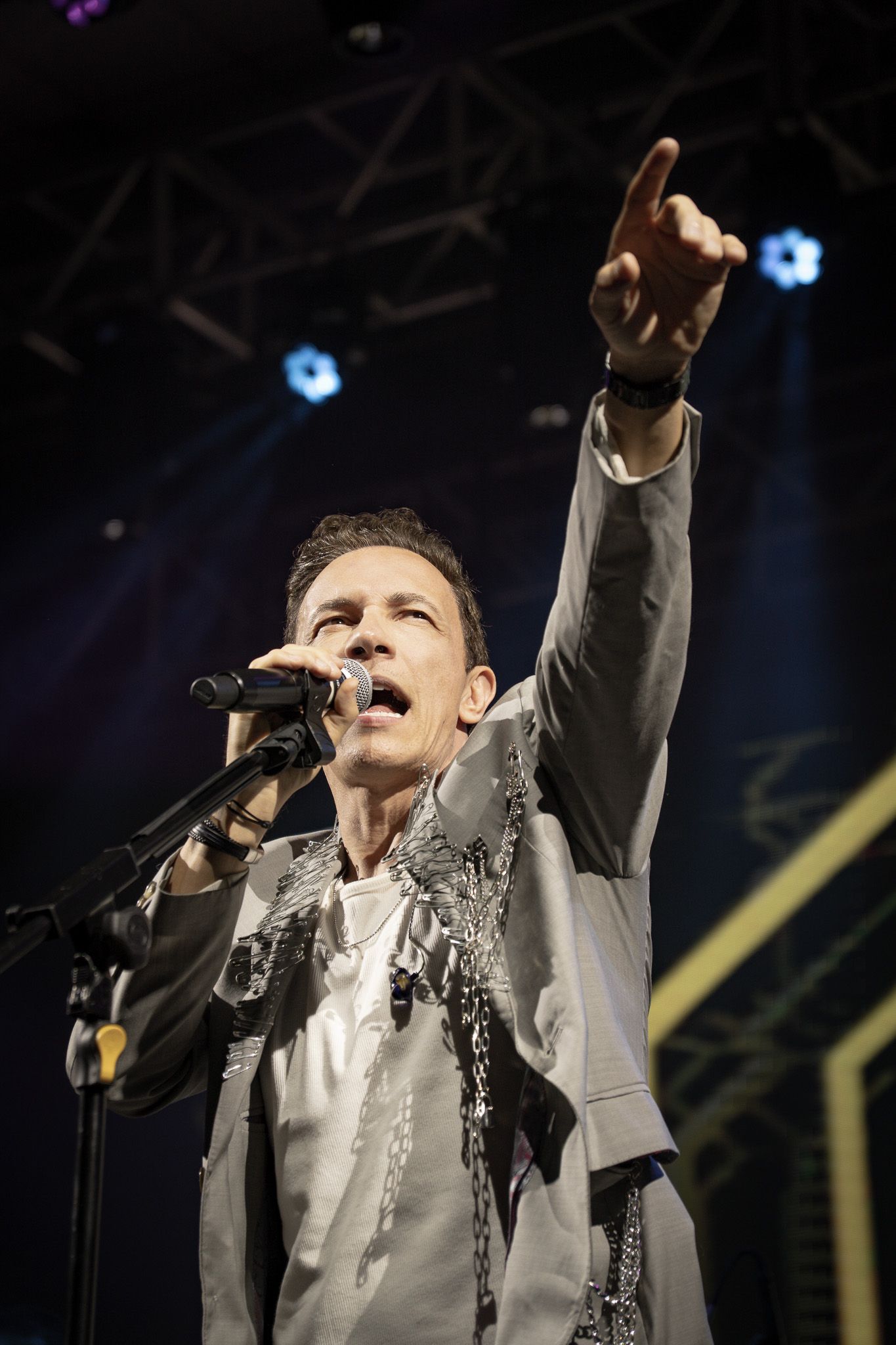 Um artista no palco cantando, apontando para cima. Vestindo jaqueta cinza com detalhes prateados, microfone na mão.