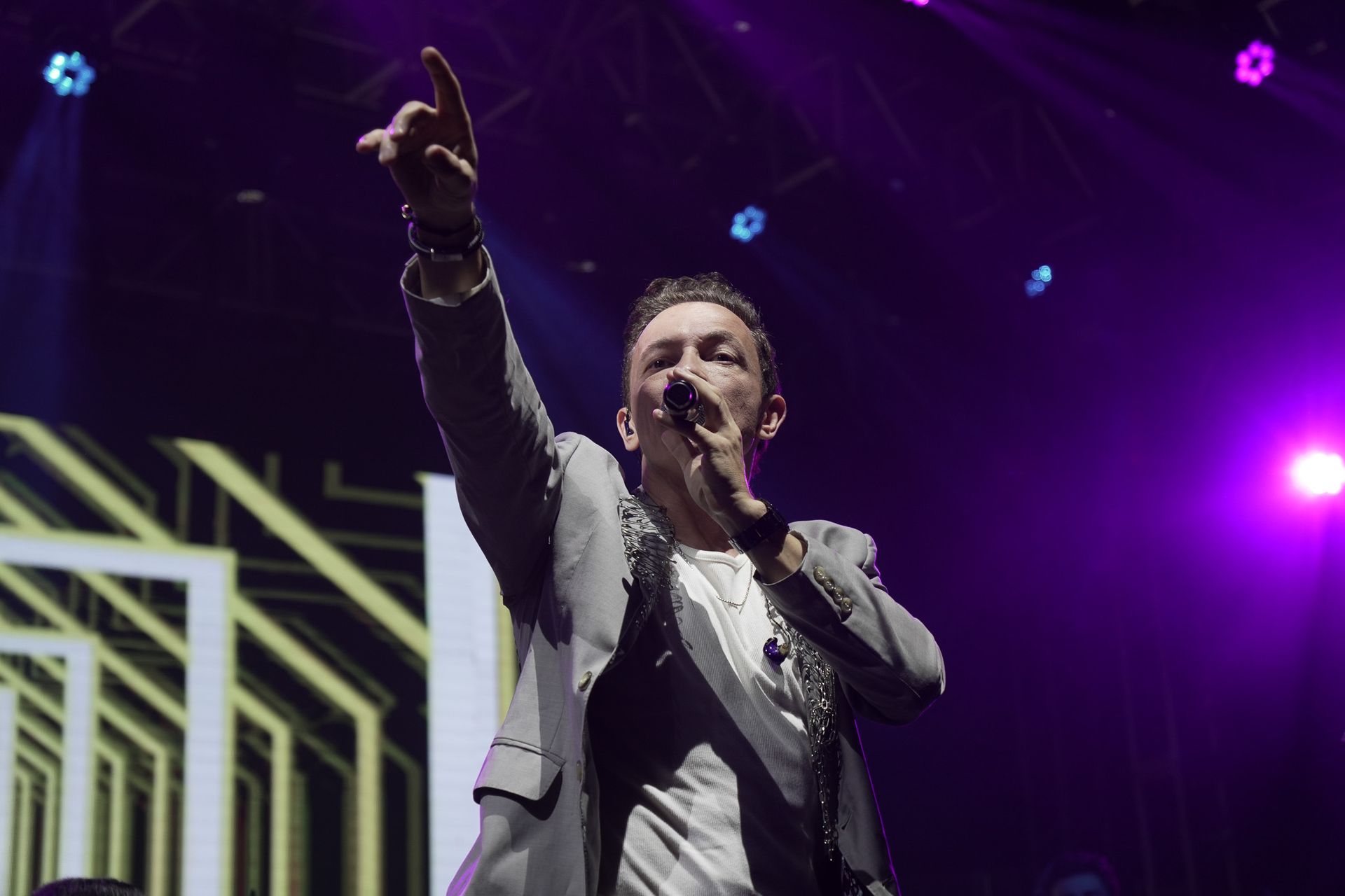 Um artista cantando em um microfone, apontando para cima com uma das mãos. Palco com iluminação roxa e dourada.