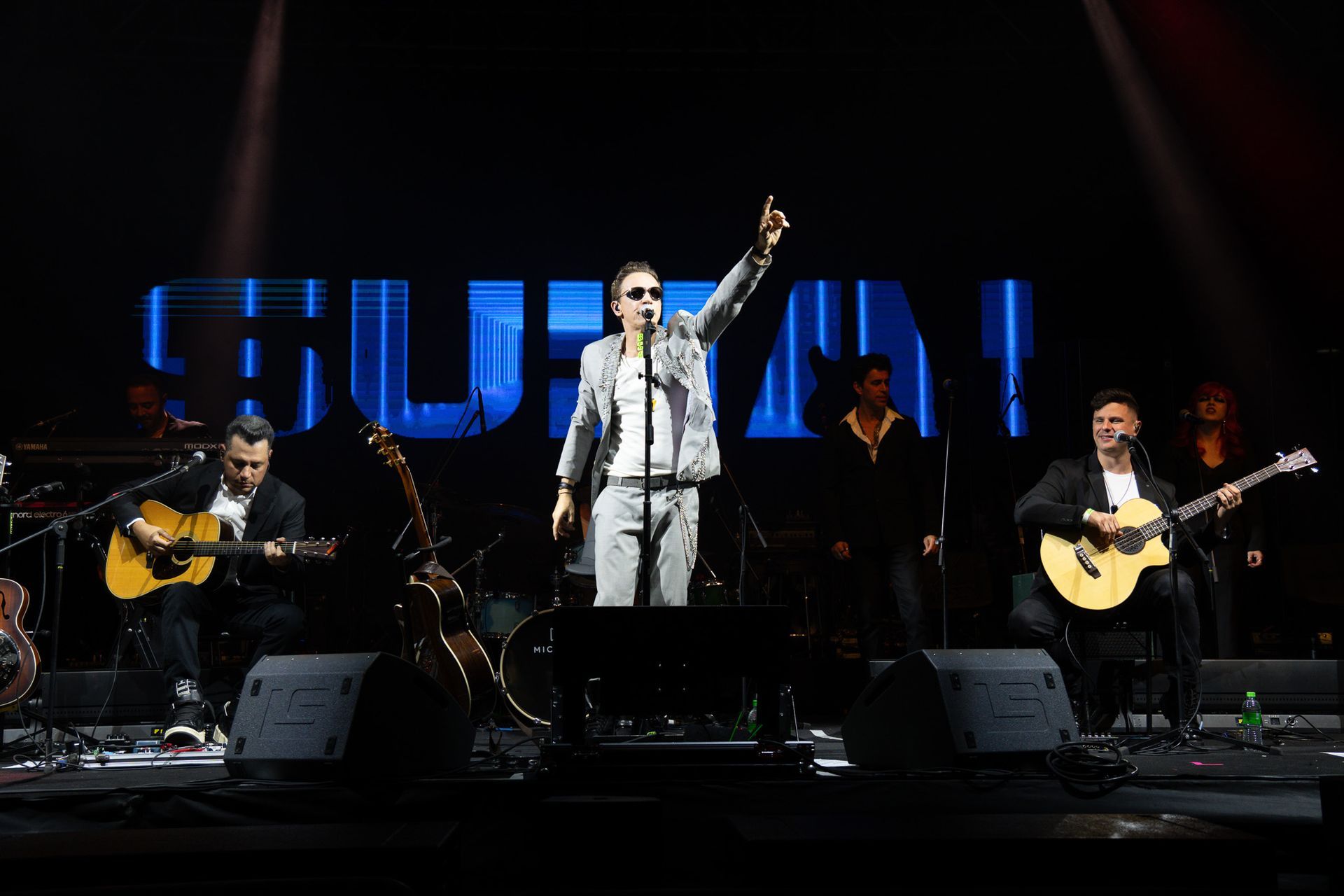 Vocalista no palco, apontando com a banda. Palco iluminado com o nome da banda 