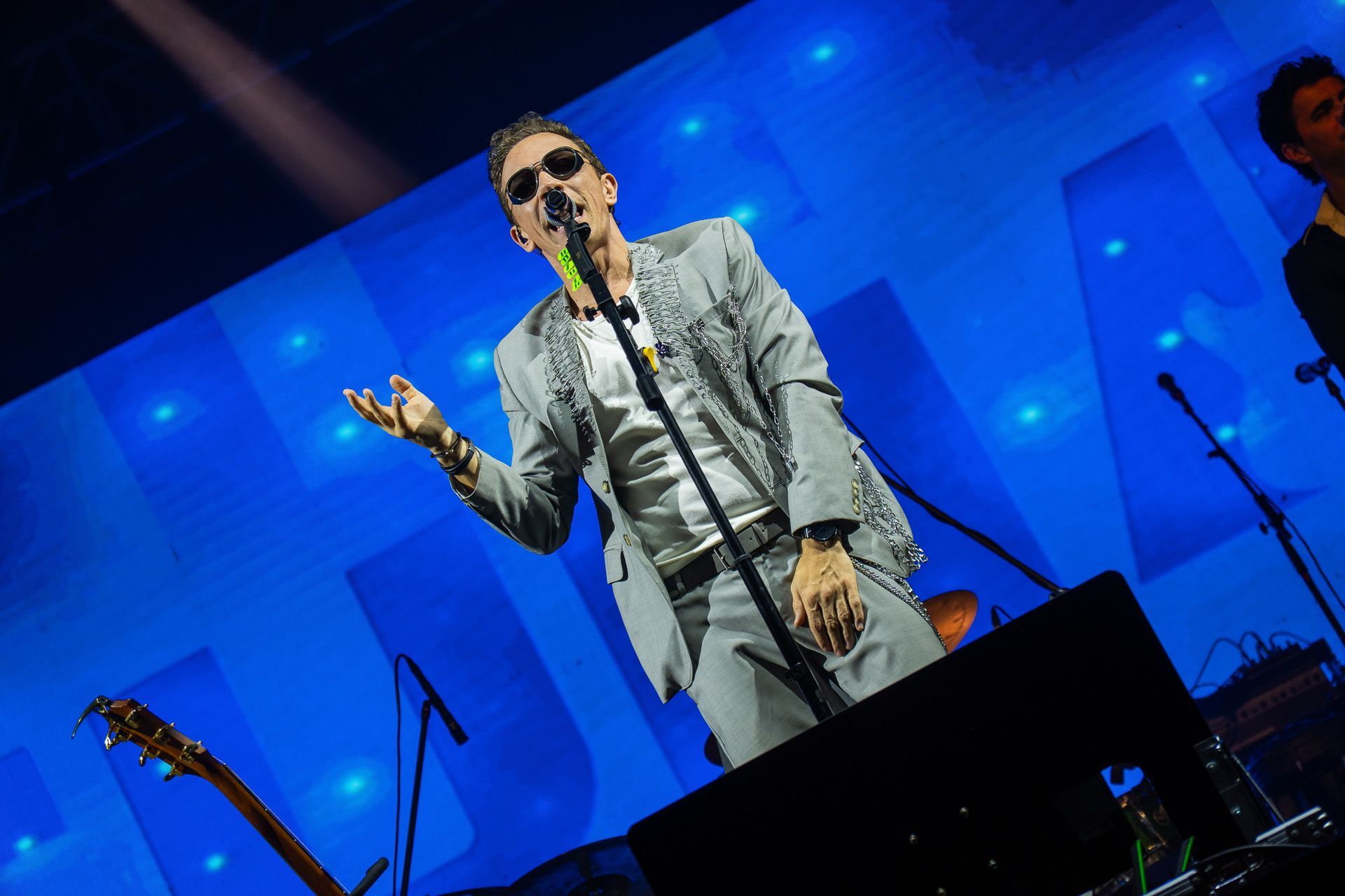 Um artista de óculos escuros e terno claro cantando no palco com fundo azul e luzes.