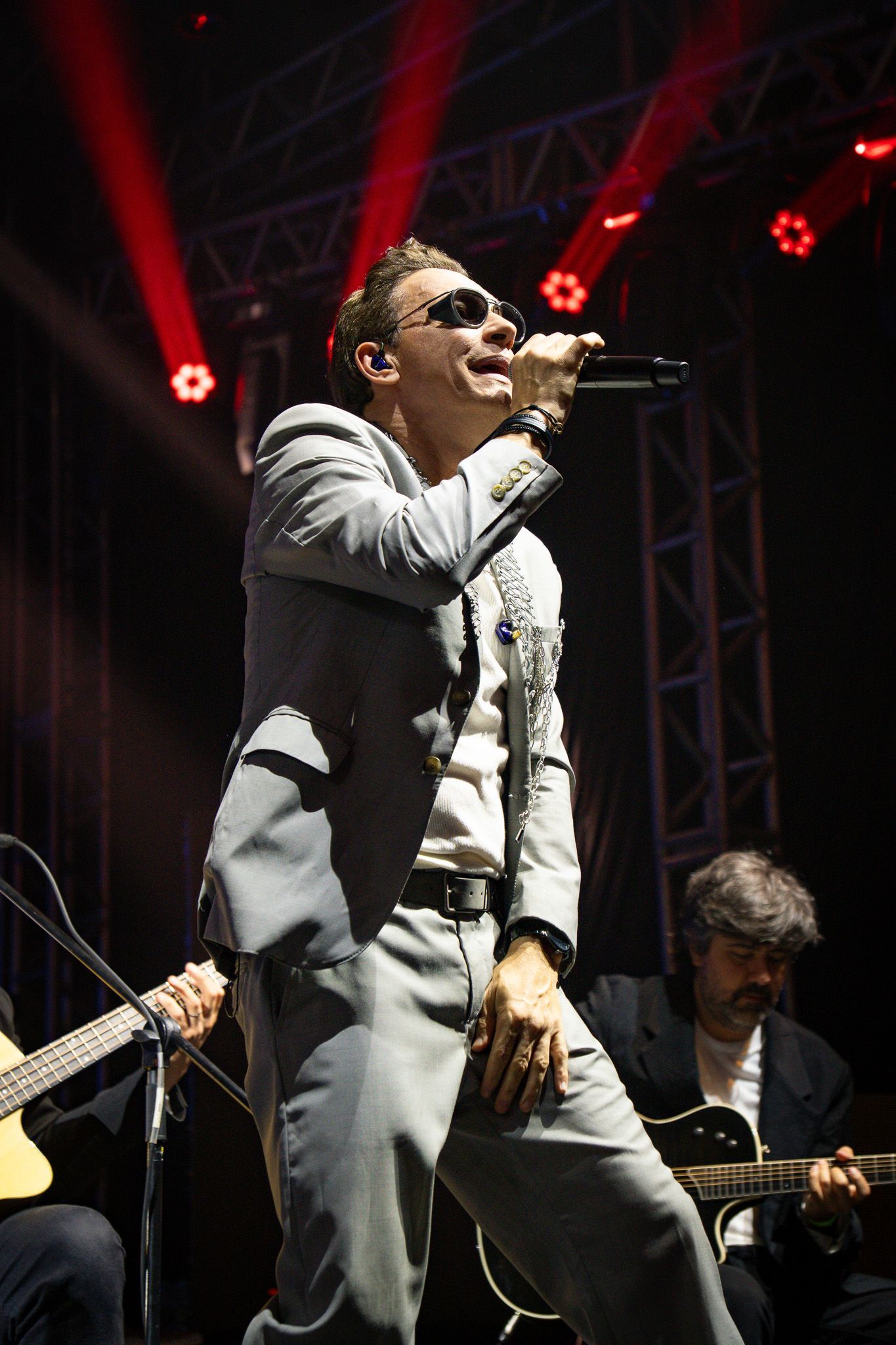 Um artista cantando ao microfone no palco, usando óculos escuros e um terno cinza. Integrantes da banda tocam guitarra ao fundo.