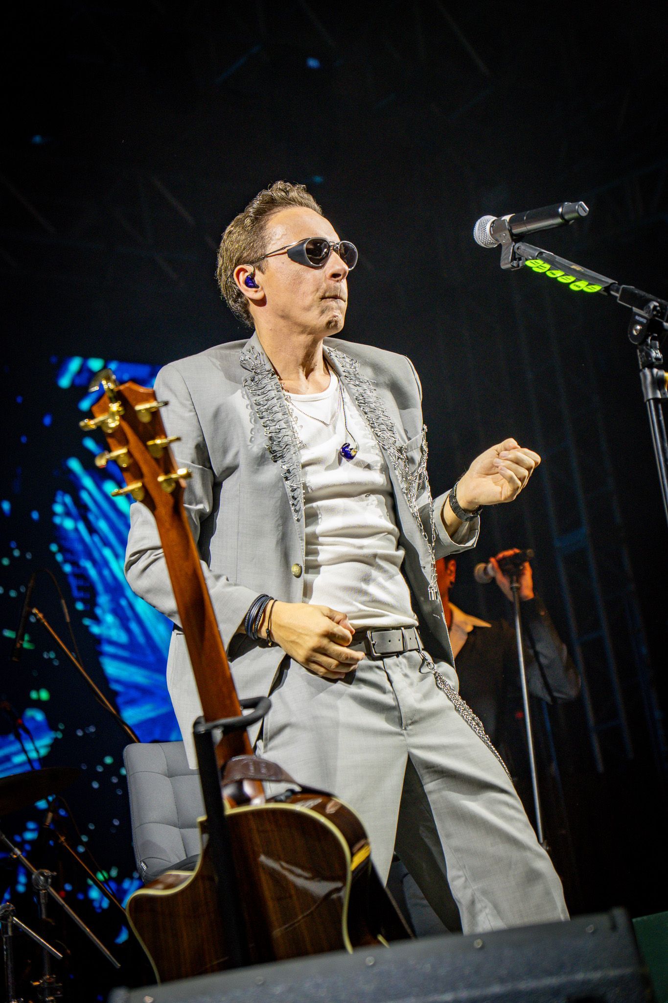 Um artista de terno cinza e óculos escuros, segurando um violão, cantando ao microfone no palco.