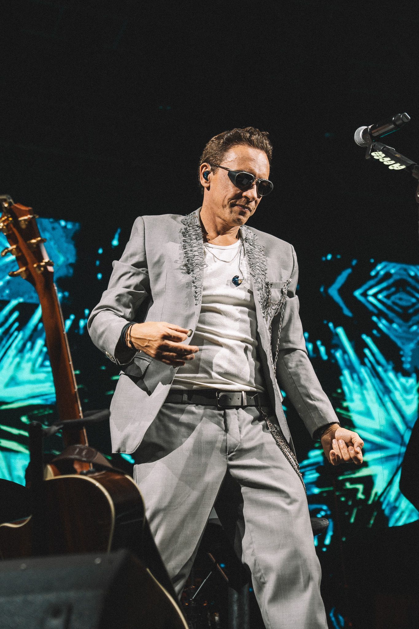 Um artista de terno cinza dançando no palco, guitarra em primeiro plano, fundo azul.