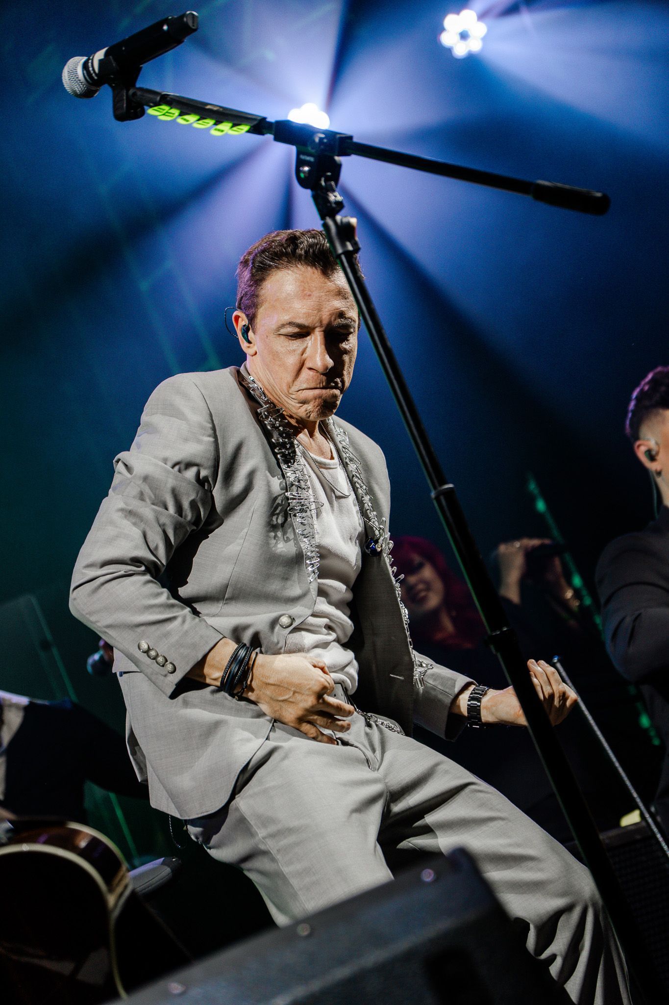 Um artista de terno cinza cantando no palco, inclinado para a frente, segurando o microfone, em um cenário de show com iluminação.