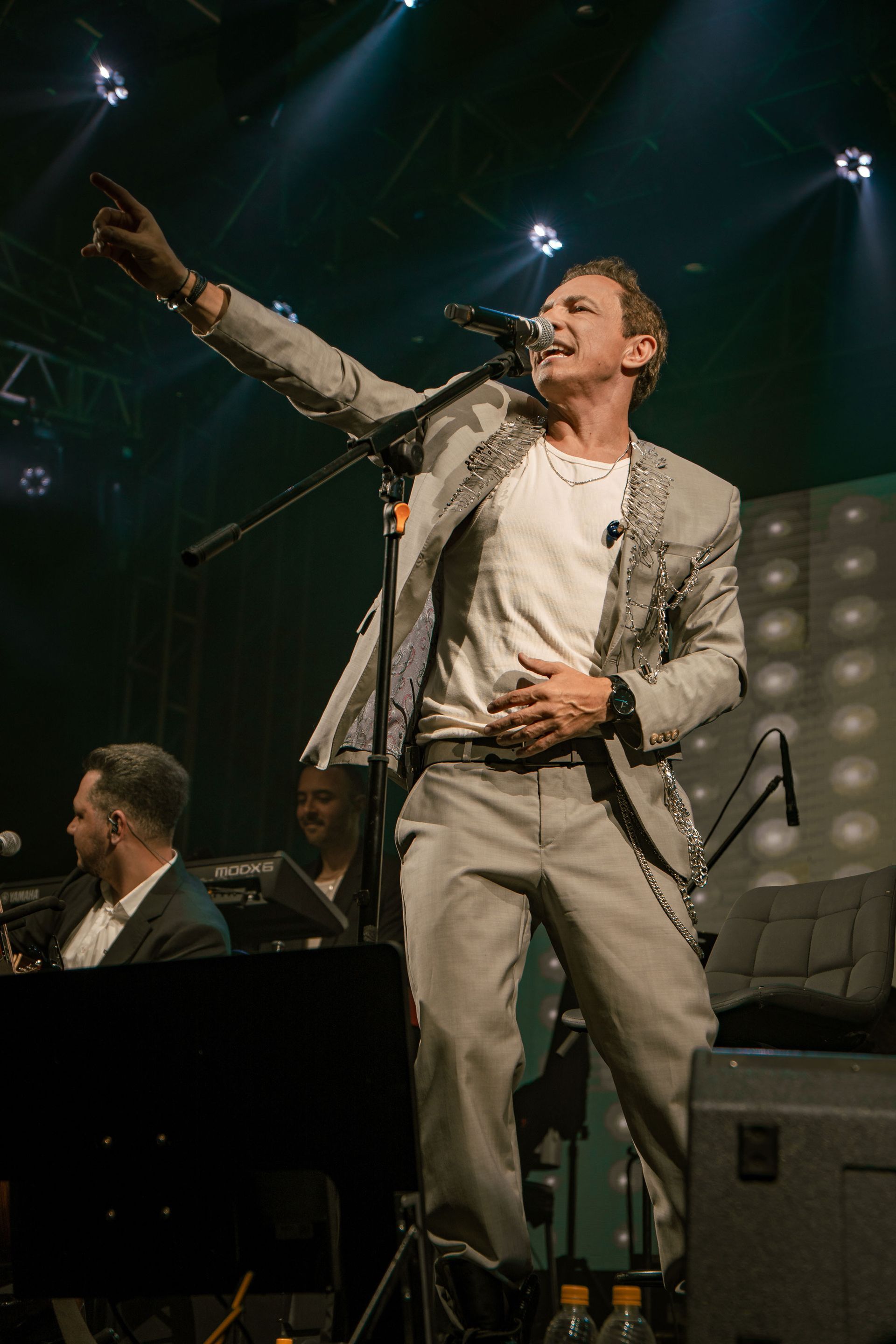Homem de terno cinza cantando no palco, braço levantado, apontando, banda ao fundo.