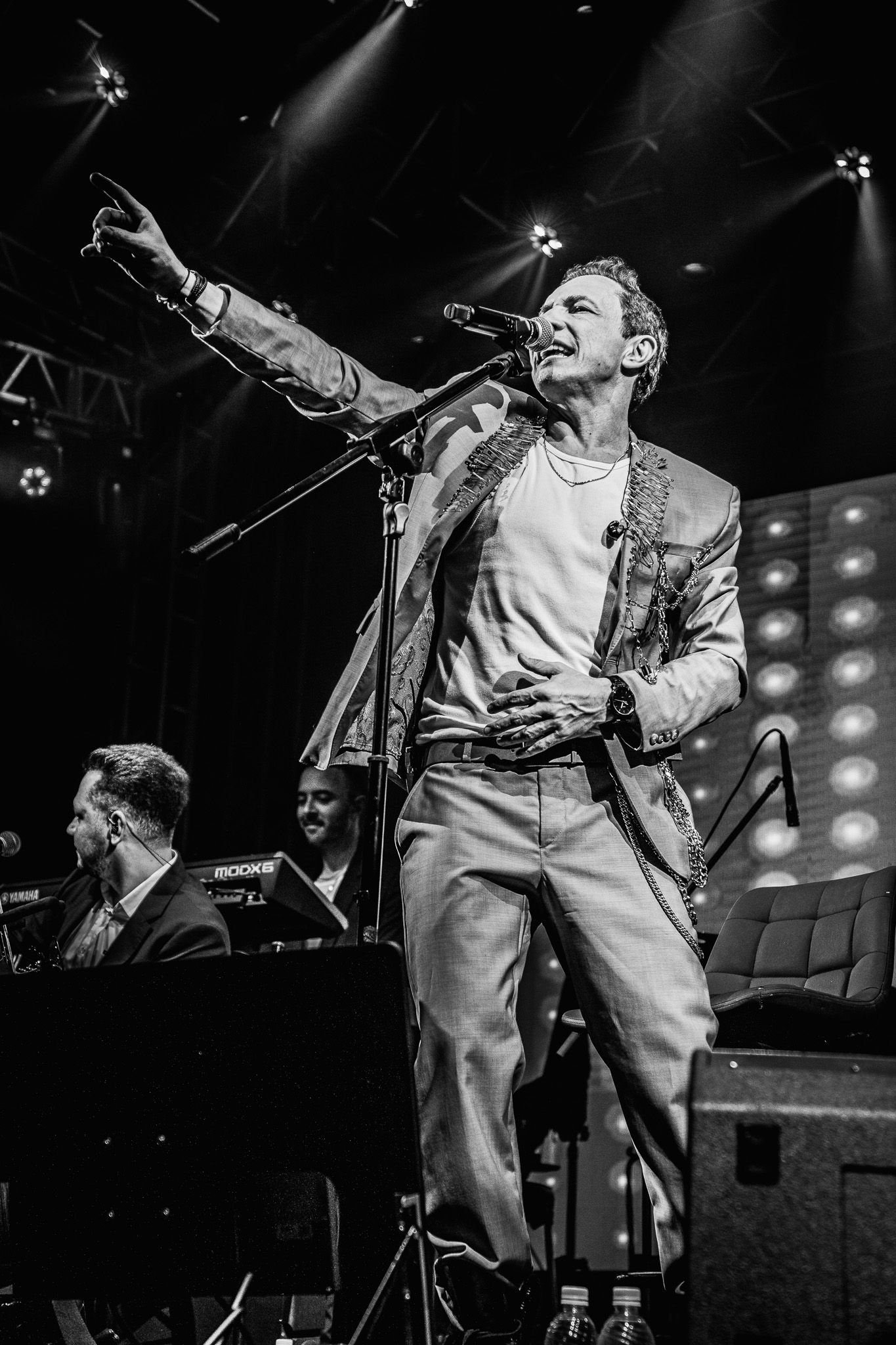 Um artista se apresentando no palco, microfone na mão, apontando para cima, com a banda ao fundo.