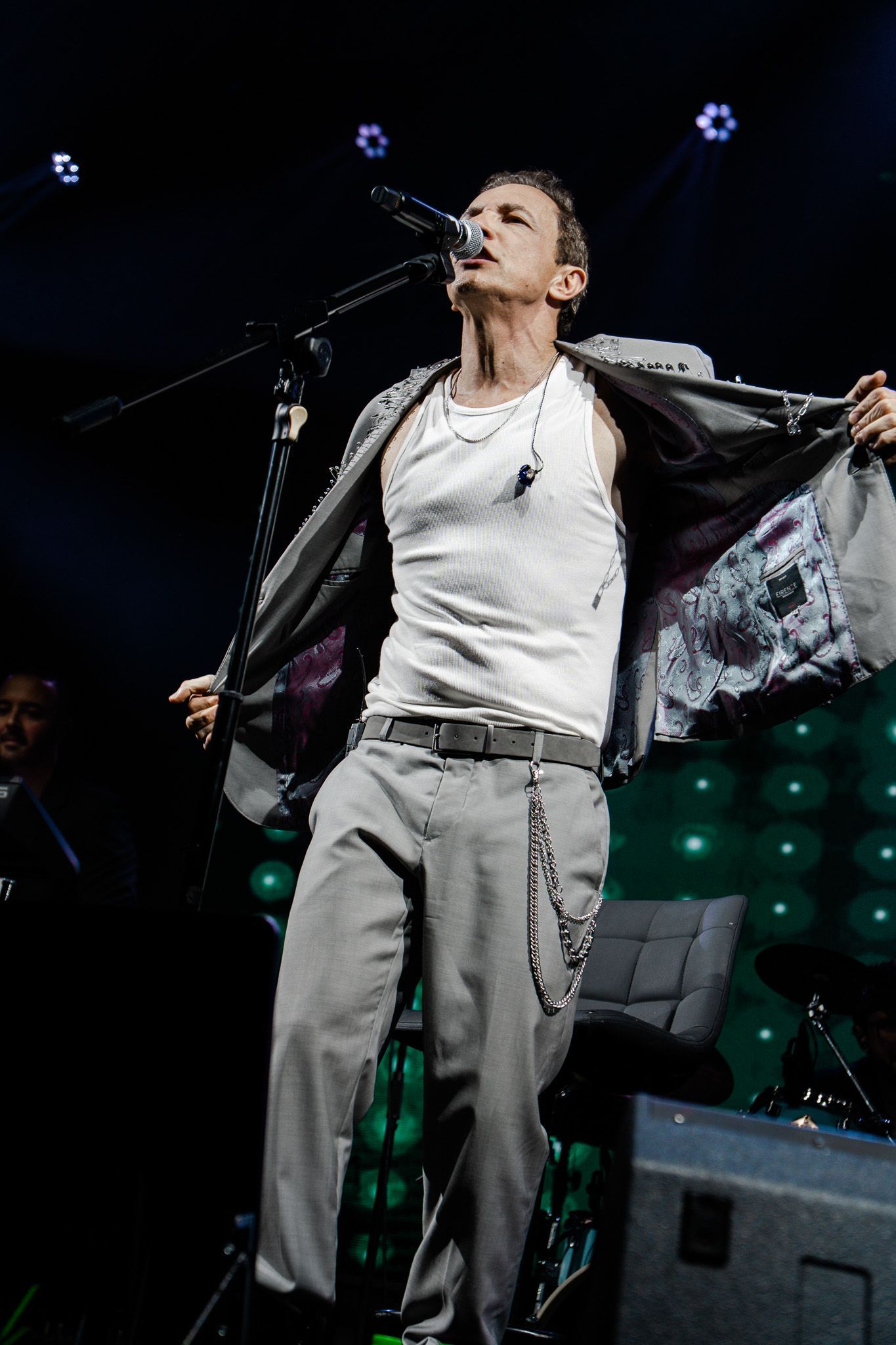 Um artista no palco, cantando, levantando o blazer, regata branca, calça cinza, microfone. Luzes verdes ao fundo.