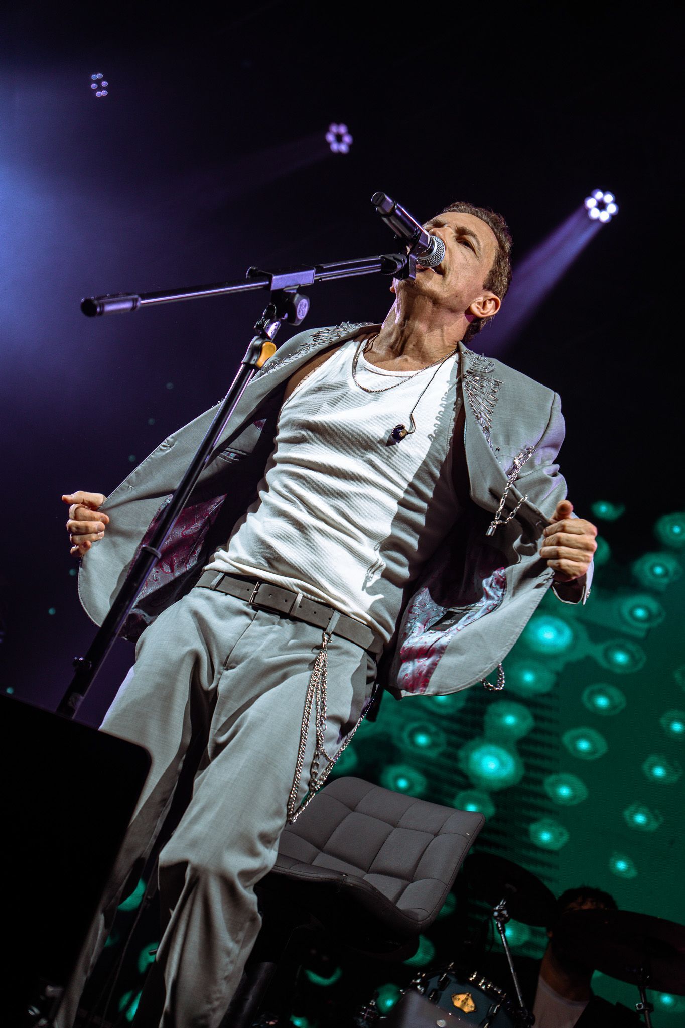 Um artista no palco cantando, vestindo terno prateado e camisa branca. Recostado, com o microfone na mão.