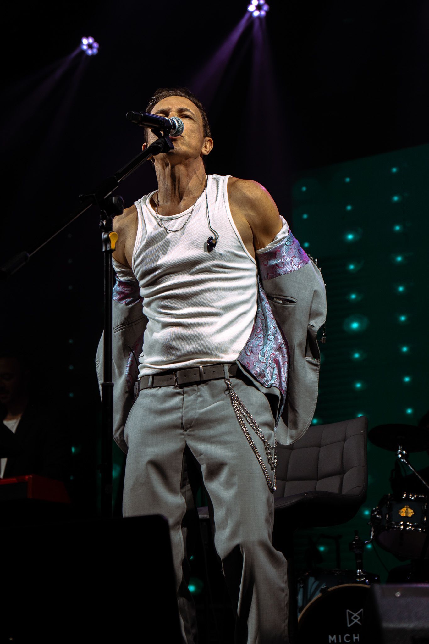 Cantor no palco, vestindo uma regata branca e calças claras, cantando ao microfone. Palco escuro com holofotes.