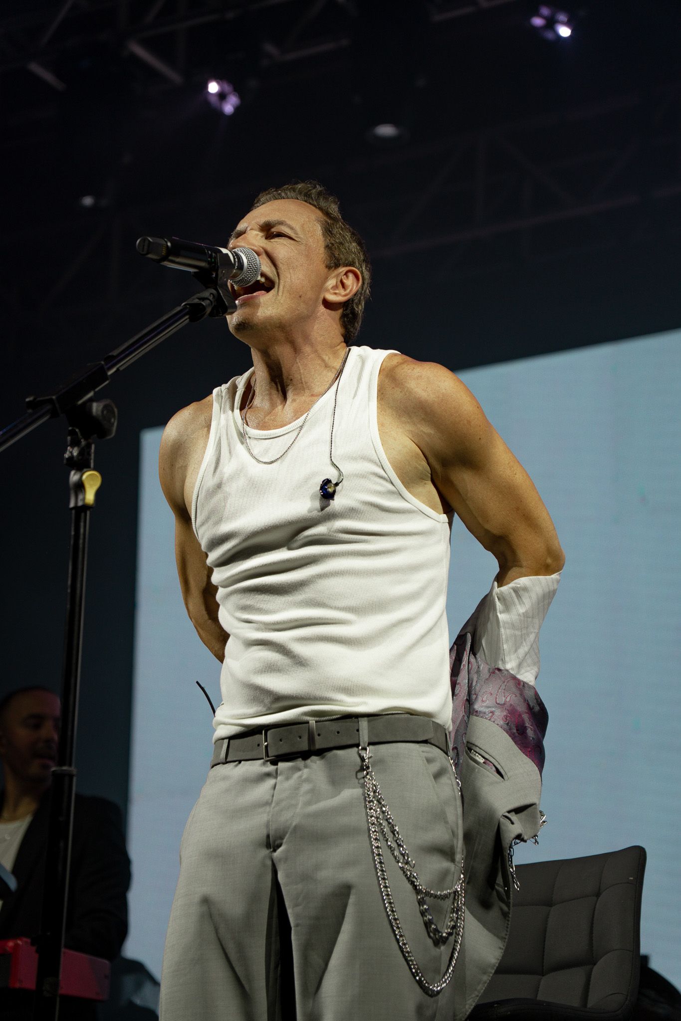 Cantor no palco, vestindo uma regata branca e calças claras, cantando ao microfone. Palco escuro com holofotes.