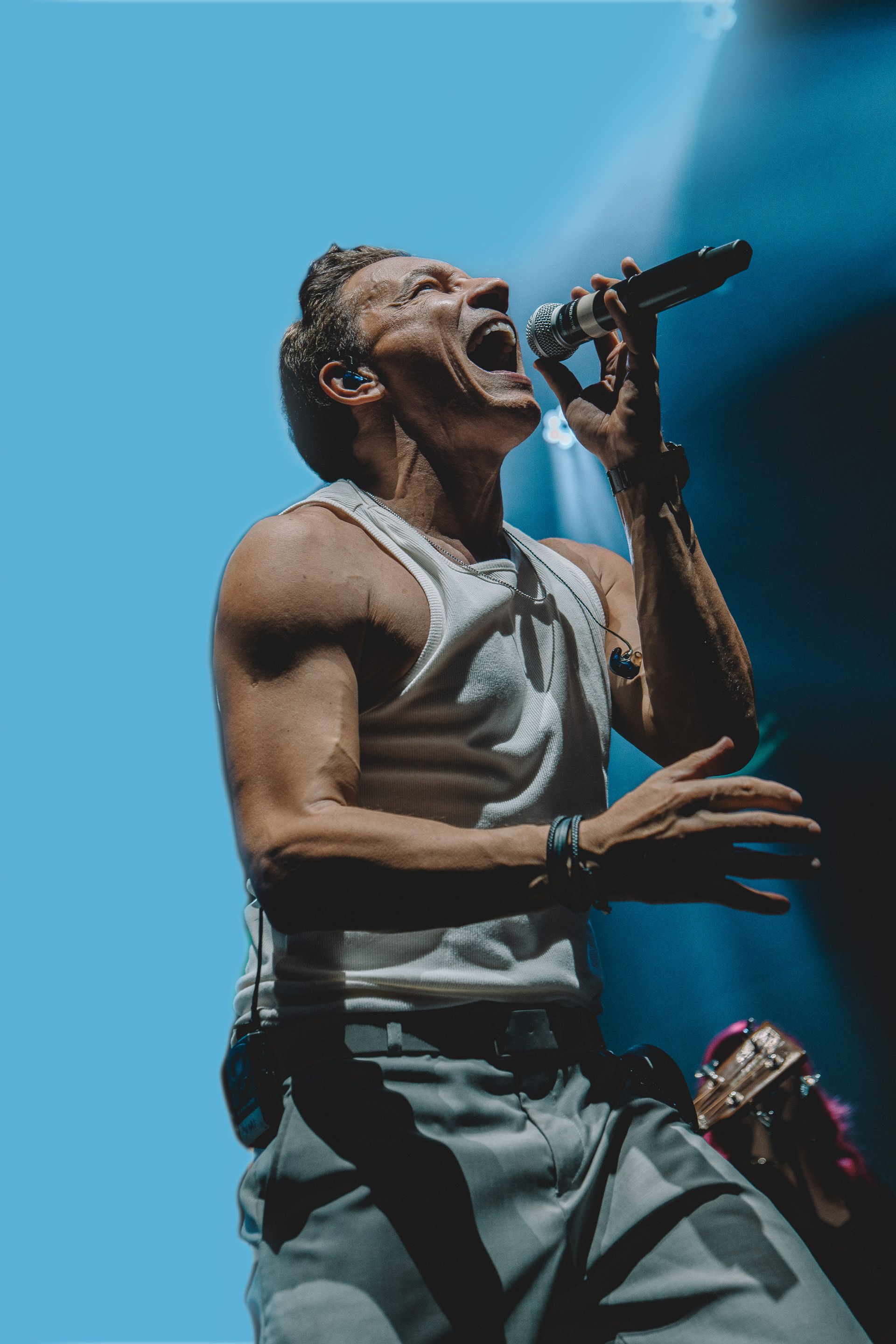 Uma pessoa vestindo uma regata branca cantando com paixão em um microfone sob luzes azuis de palco.