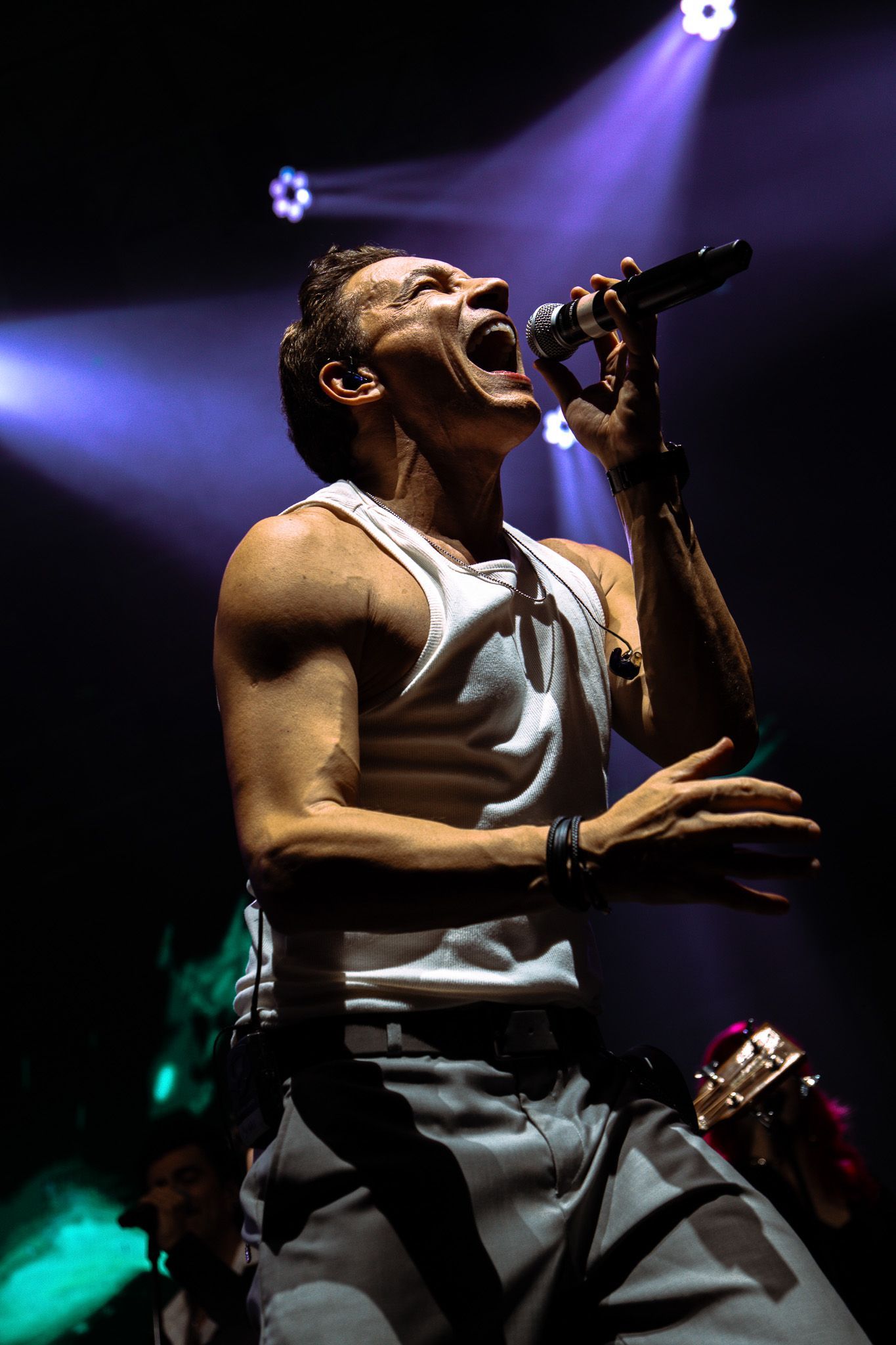 Cantor no palco, boca aberta, cantando em um microfone. Vestindo regata branca, se apresentando sob luzes de palco.