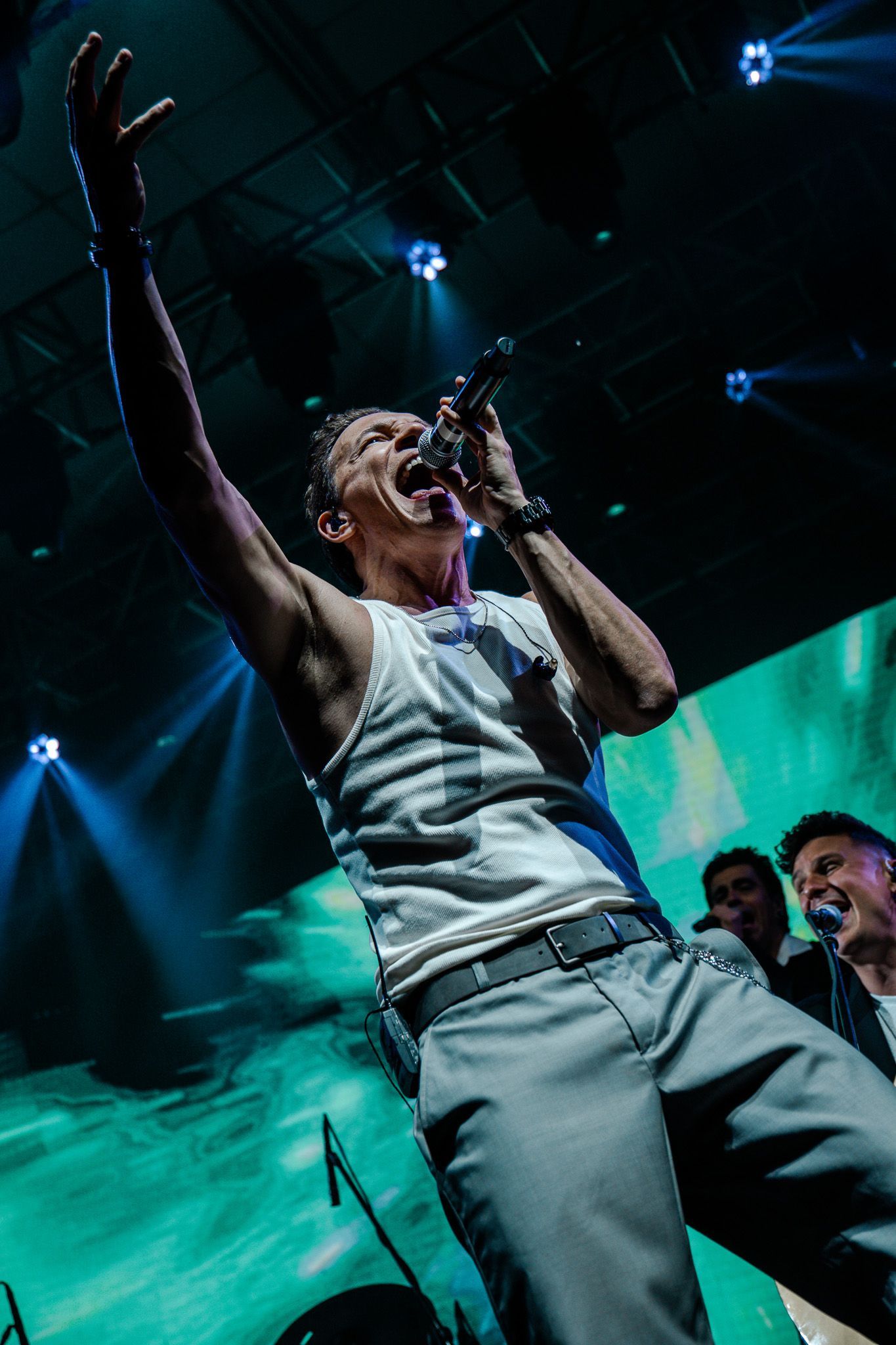 Um artista no palco, cantando com o braço levantado, vestindo uma regata branca. Outros músicos ao fundo.