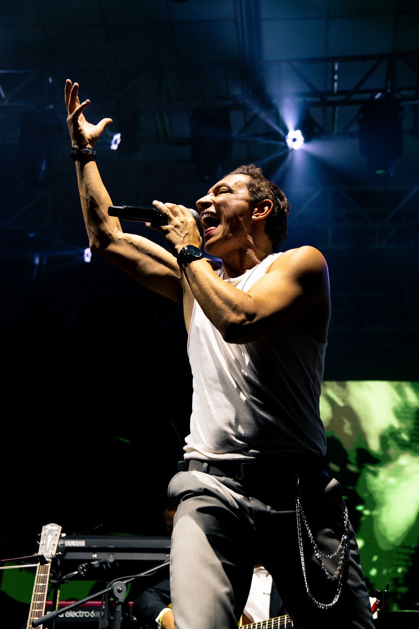 Cantor no palco, braço levantado, segurando o microfone. Regata branca, calça escura. Palco iluminado por holofote.