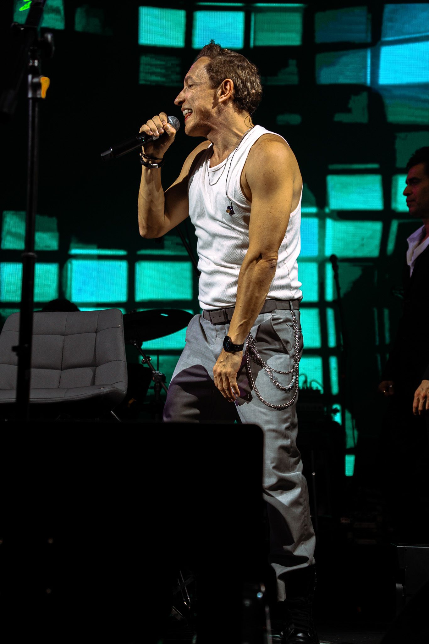 Um artista cantando ao microfone no palco, vestindo uma regata branca e calça jeans; fundo azul-petróleo.