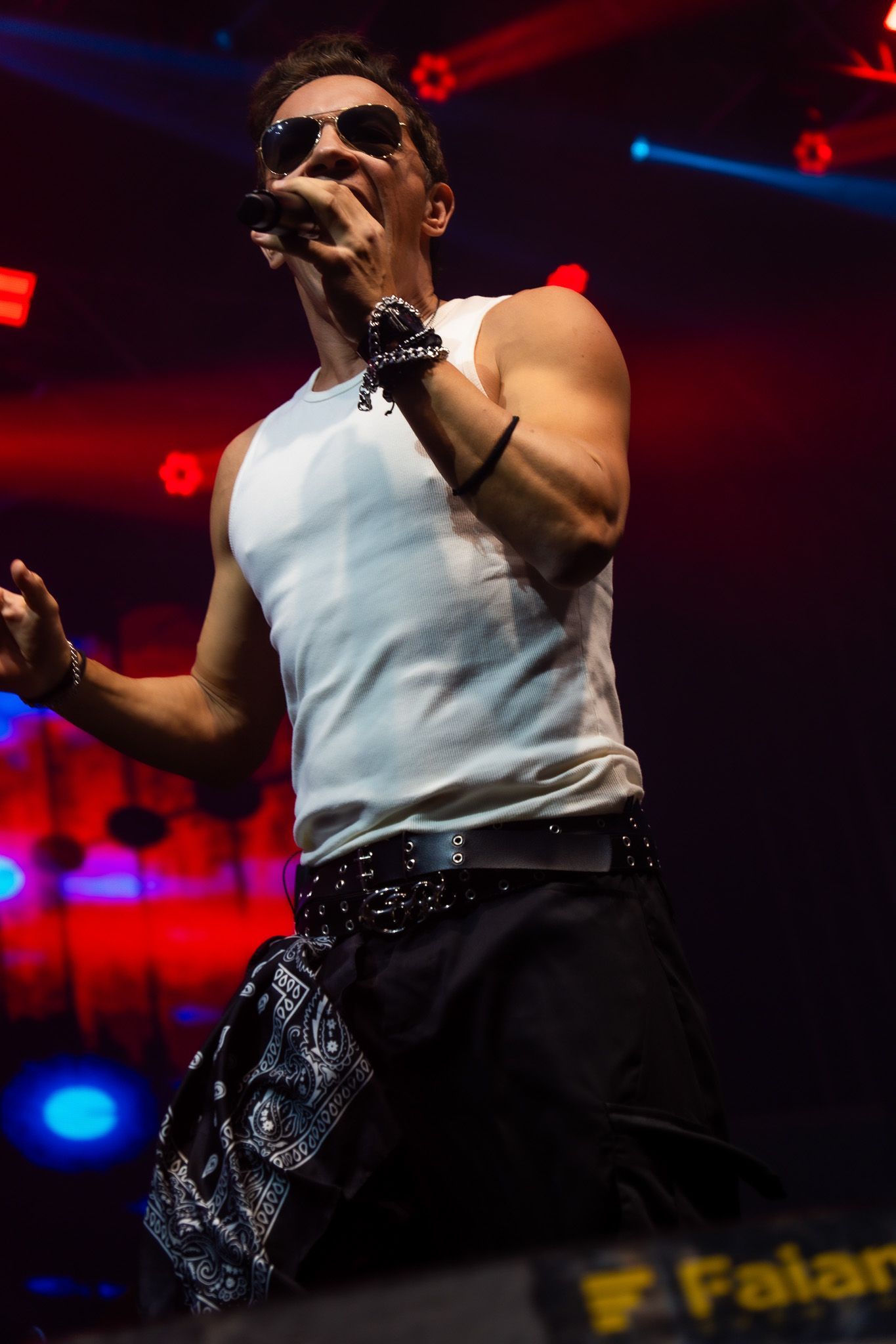 Um artista vestindo uma regata branca e óculos escuros canta em um microfone em um palco com iluminação.