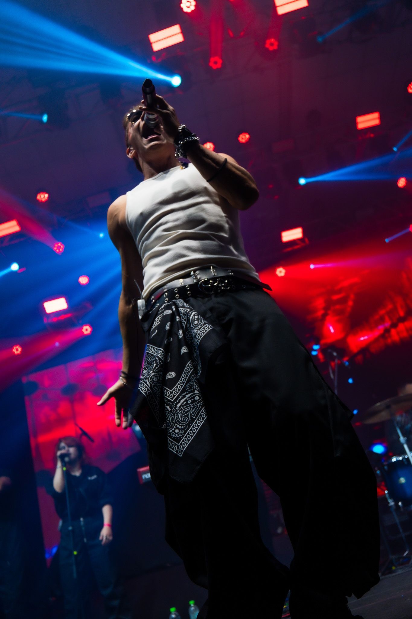 Um artista cantando ao microfone no palco, vestindo uma regata branca e calças pretas com um acessório de tecido estampado.