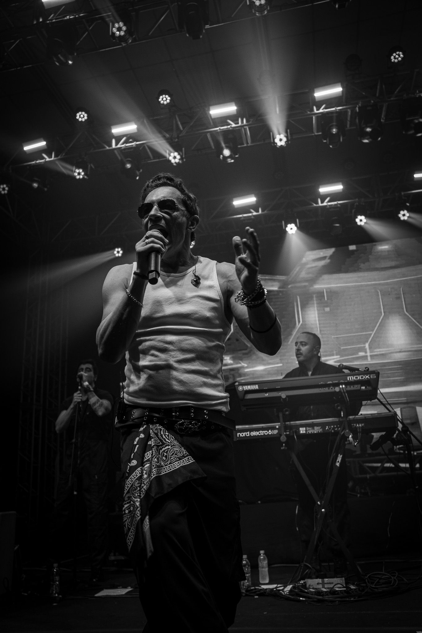 Uma pessoa canta em um microfone no palco de um show, vestindo uma regata e uma bandana, com um tecladista ao fundo.