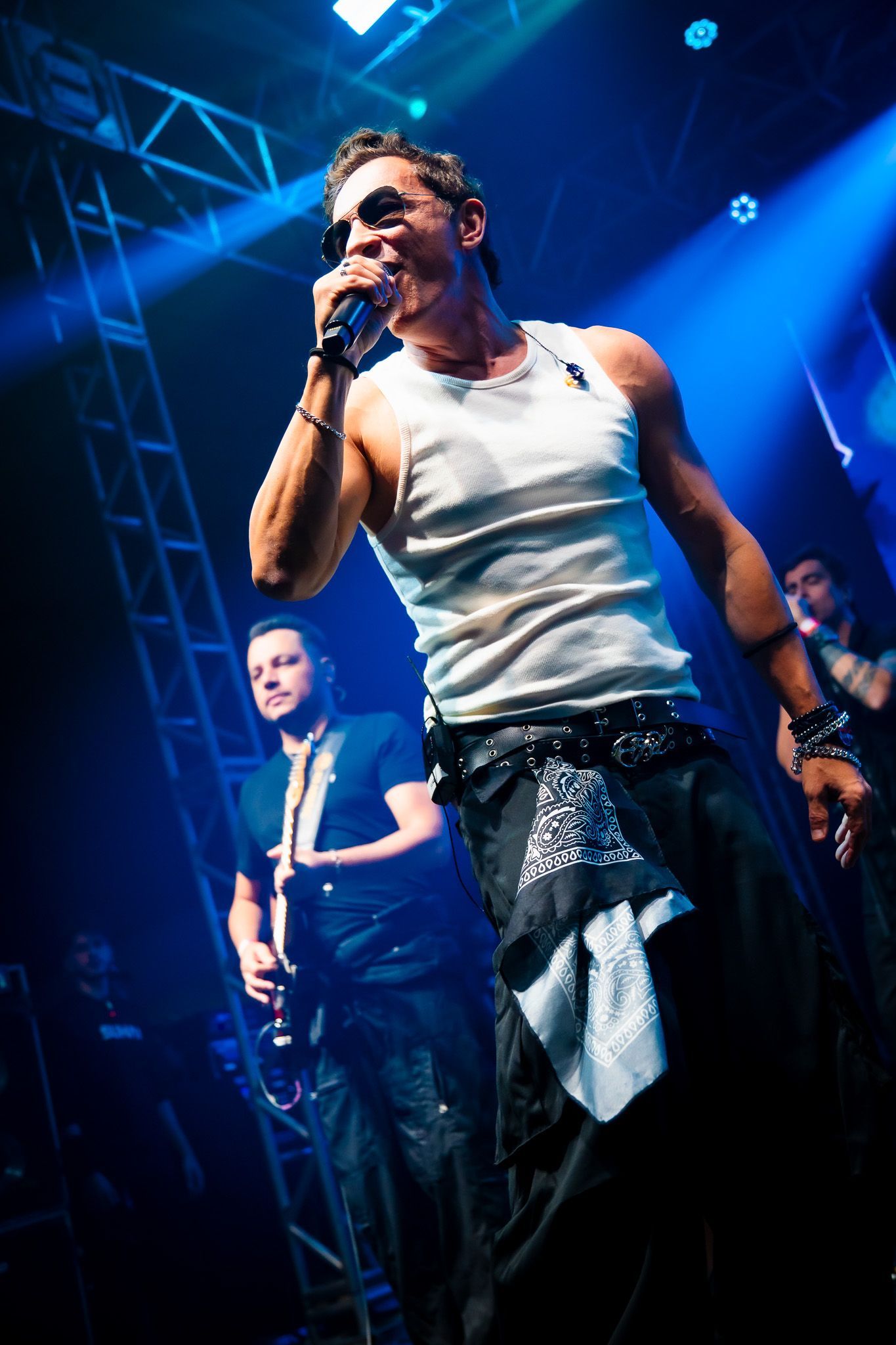 Um cantor se apresenta ao microfone no palco, iluminada por holofotes azuis, com um membro da banda tocando guitarra