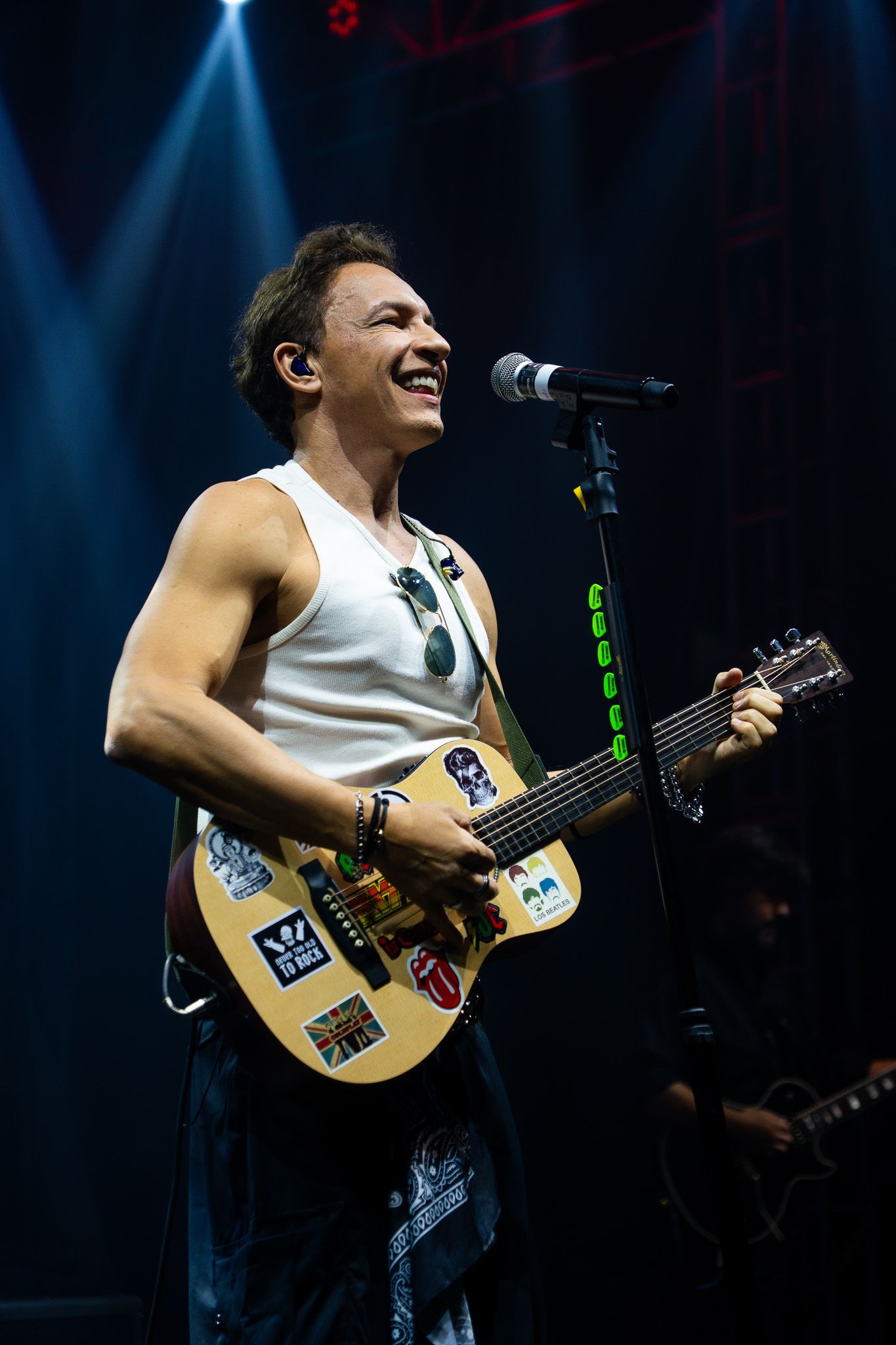 Um músico com um sorriso no rosto toca um violão decorado com adesivos durante uma apresentação no palco.