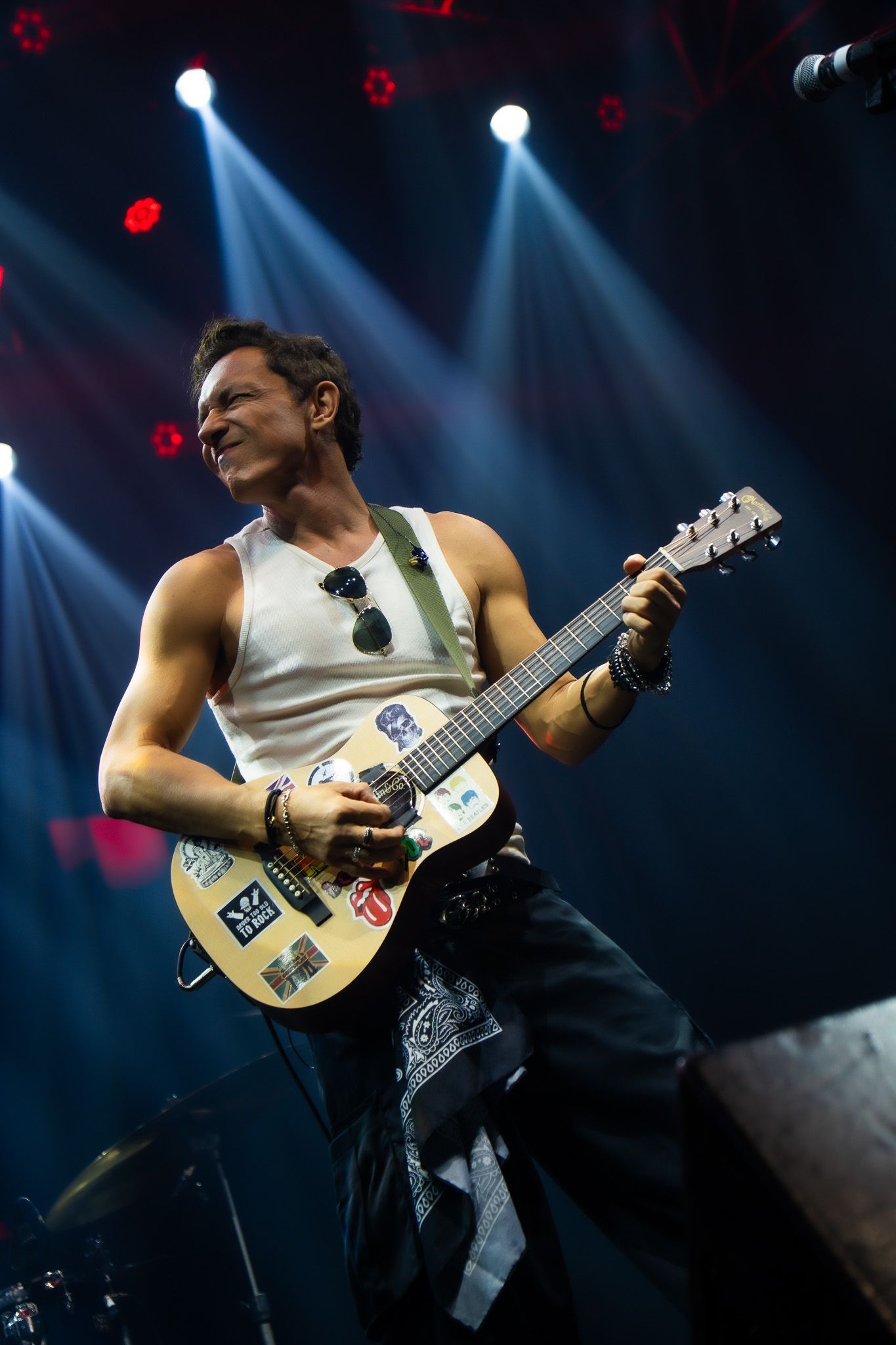 Um músico de regata branca toca uma guitarra elétrica coberta de adesivos em um palco iluminado por holofotes brilhantes.