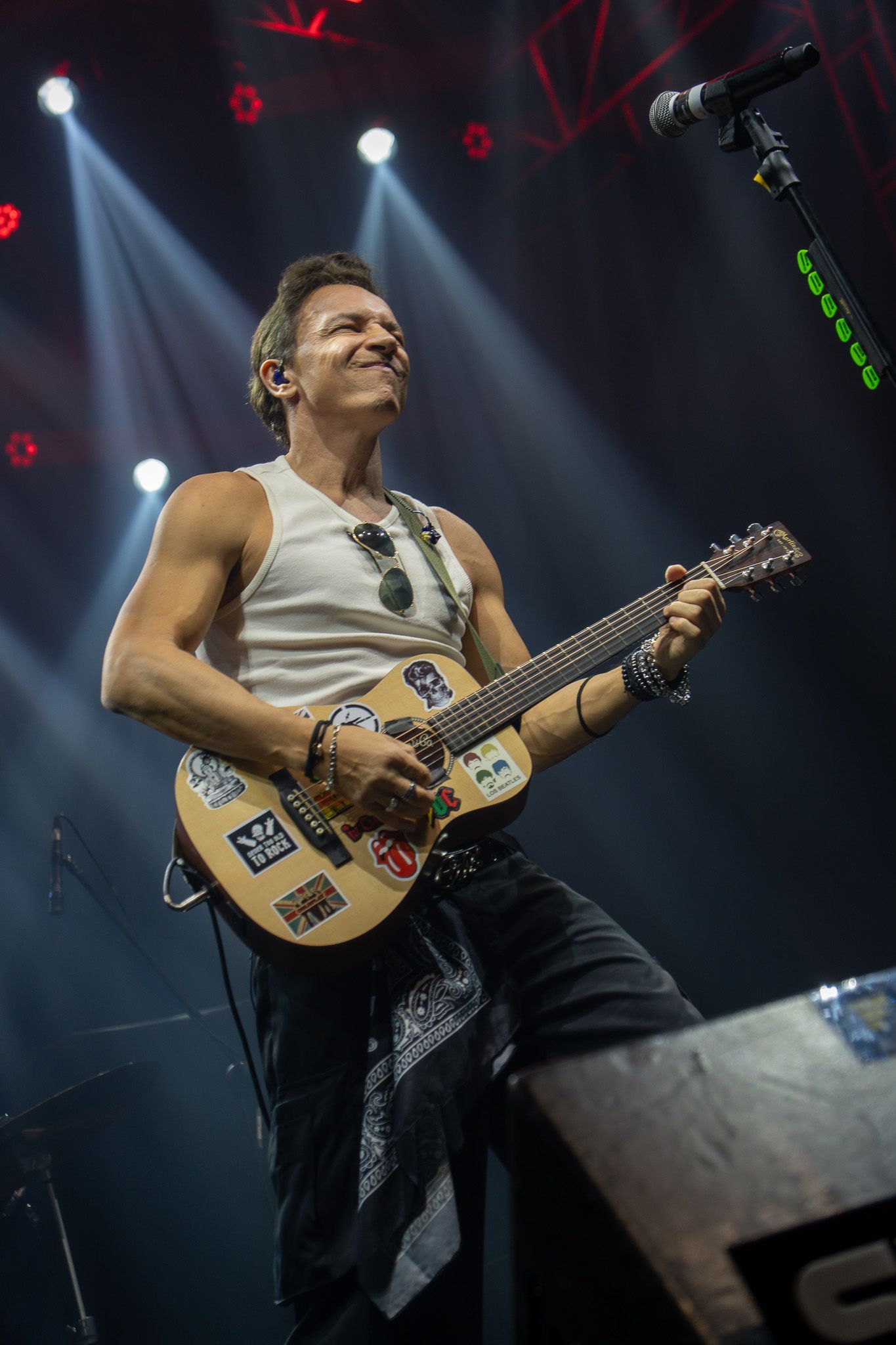 Um músico sorrindo enquanto toca um violão coberto de adesivos em um palco iluminado por holofotes brilhantes.