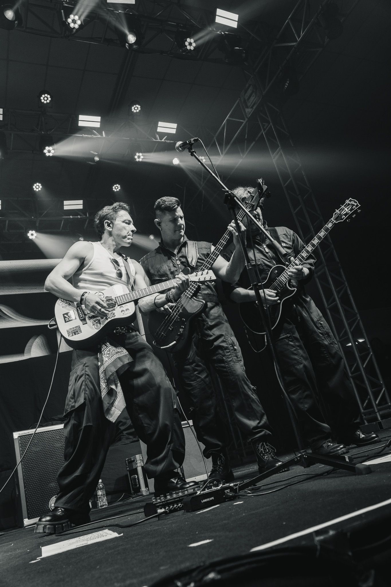 Três músicos tocando guitarras elétricas e se apresentando em um palco de concertos bem iluminado.