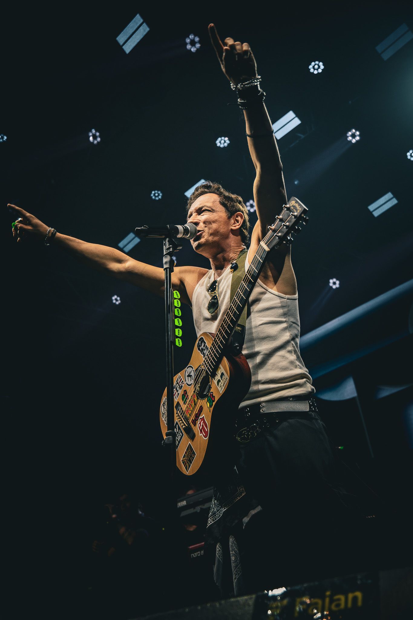 Um músico de regata branca se apresenta no palco, tocando uma guitarra elétrica estampada com um braço levantado.