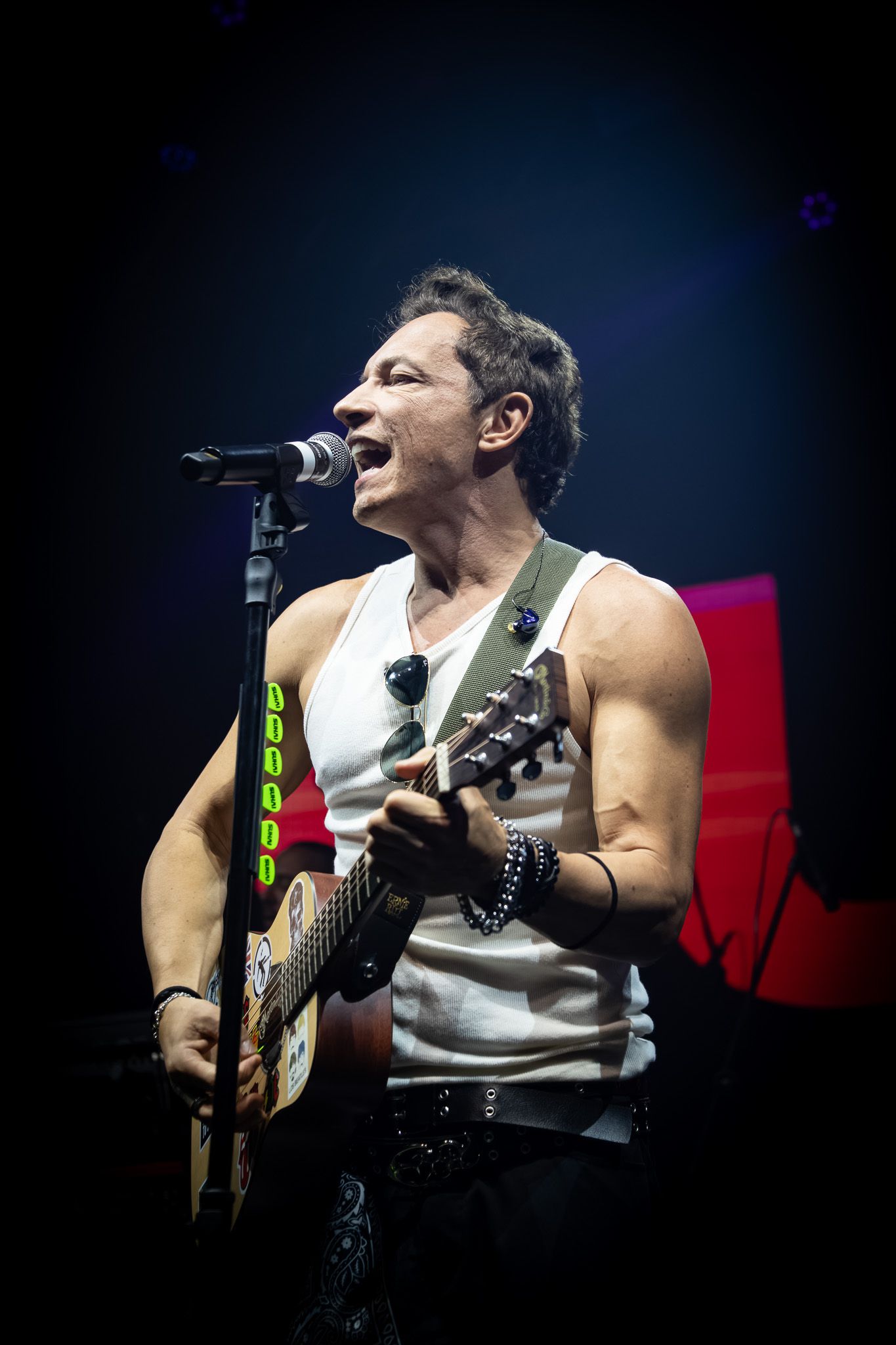 Uma pessoa vestindo uma regata branca toca violão e canta ao microfone em um palco com fundo escuro.