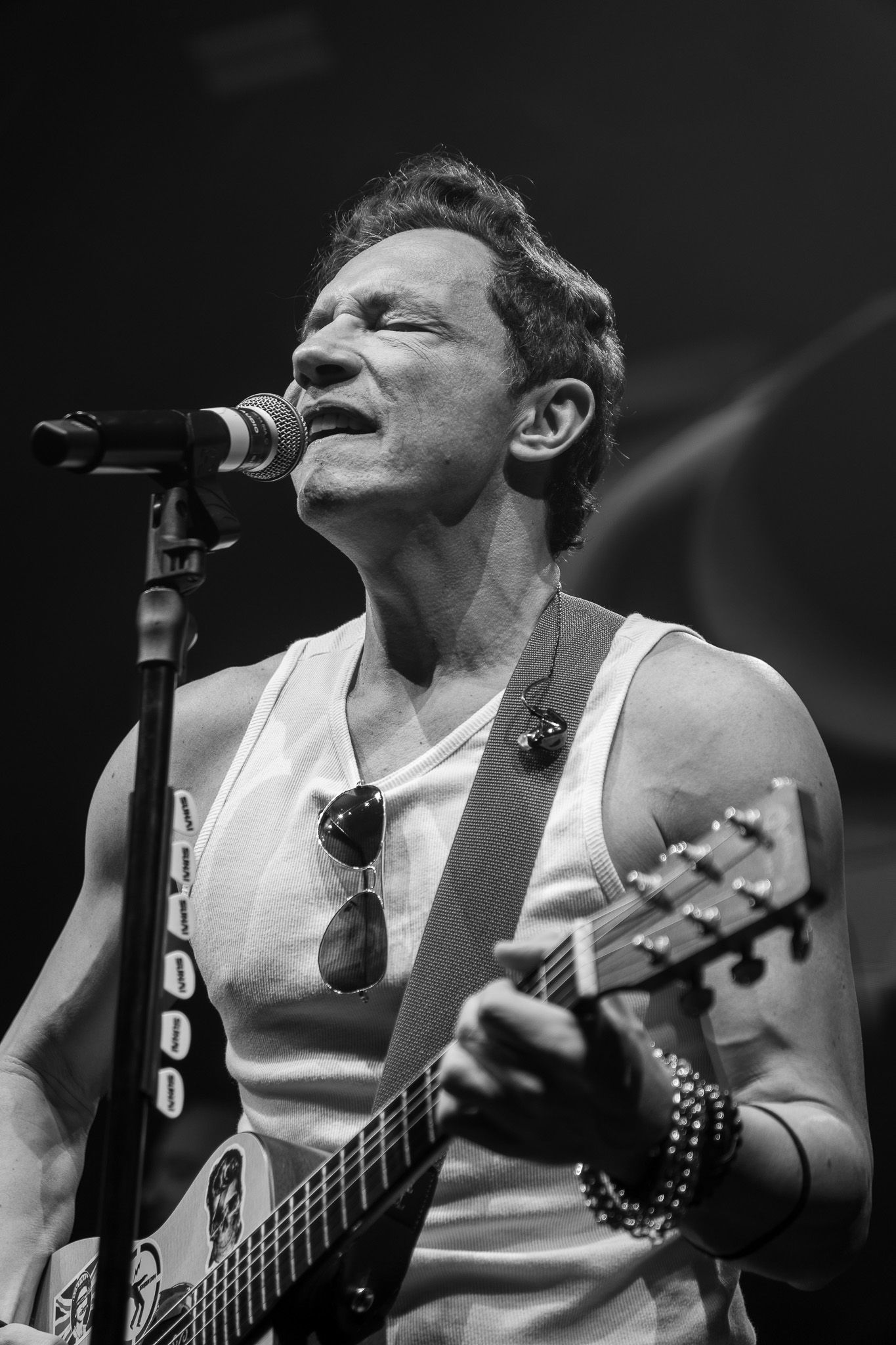 Fotografia em preto e branco de um músico de regata cantando ao microfone enquanto toca guitarra elétrica no palco.