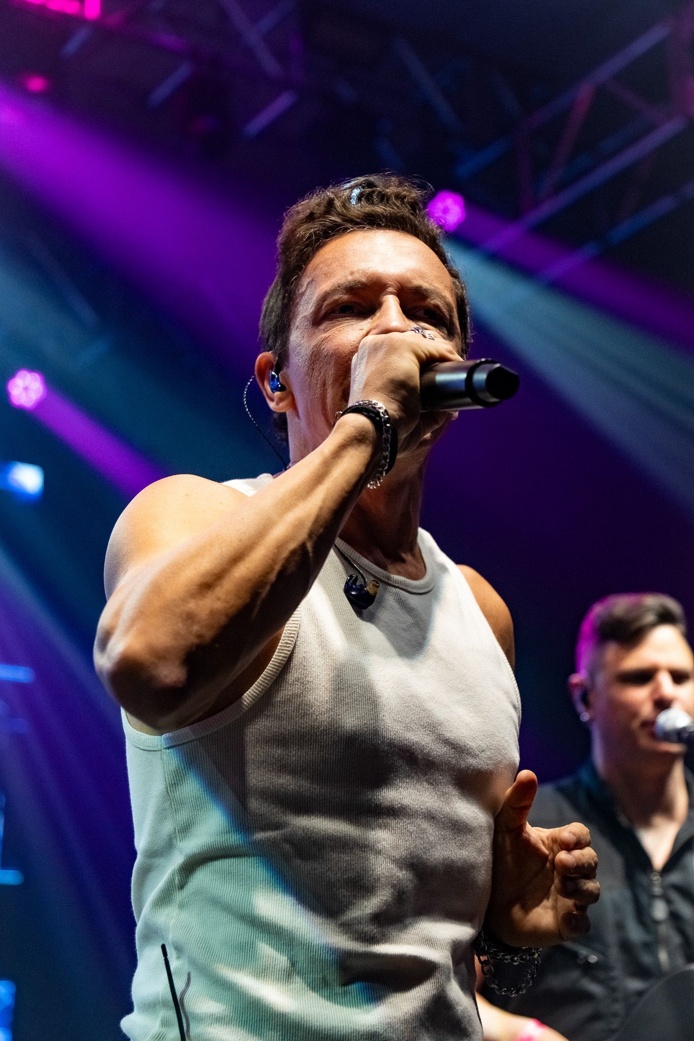 Um artista cantando em um microfone em um palco com iluminação roxa e outro músico ao fundo.