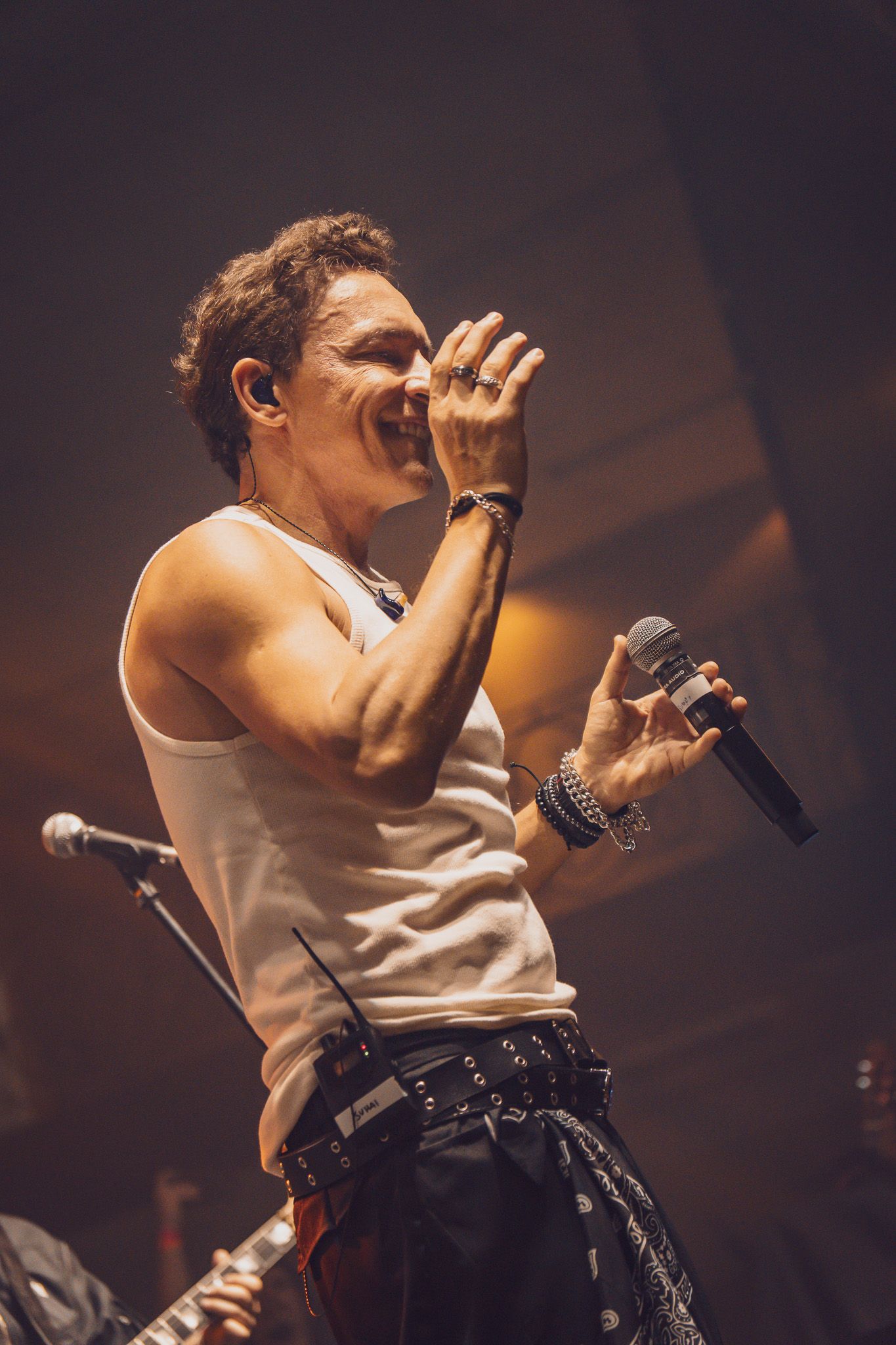 Um músico sorridente, vestindo uma regata branca, se apresenta no palco, segurando um microfone 